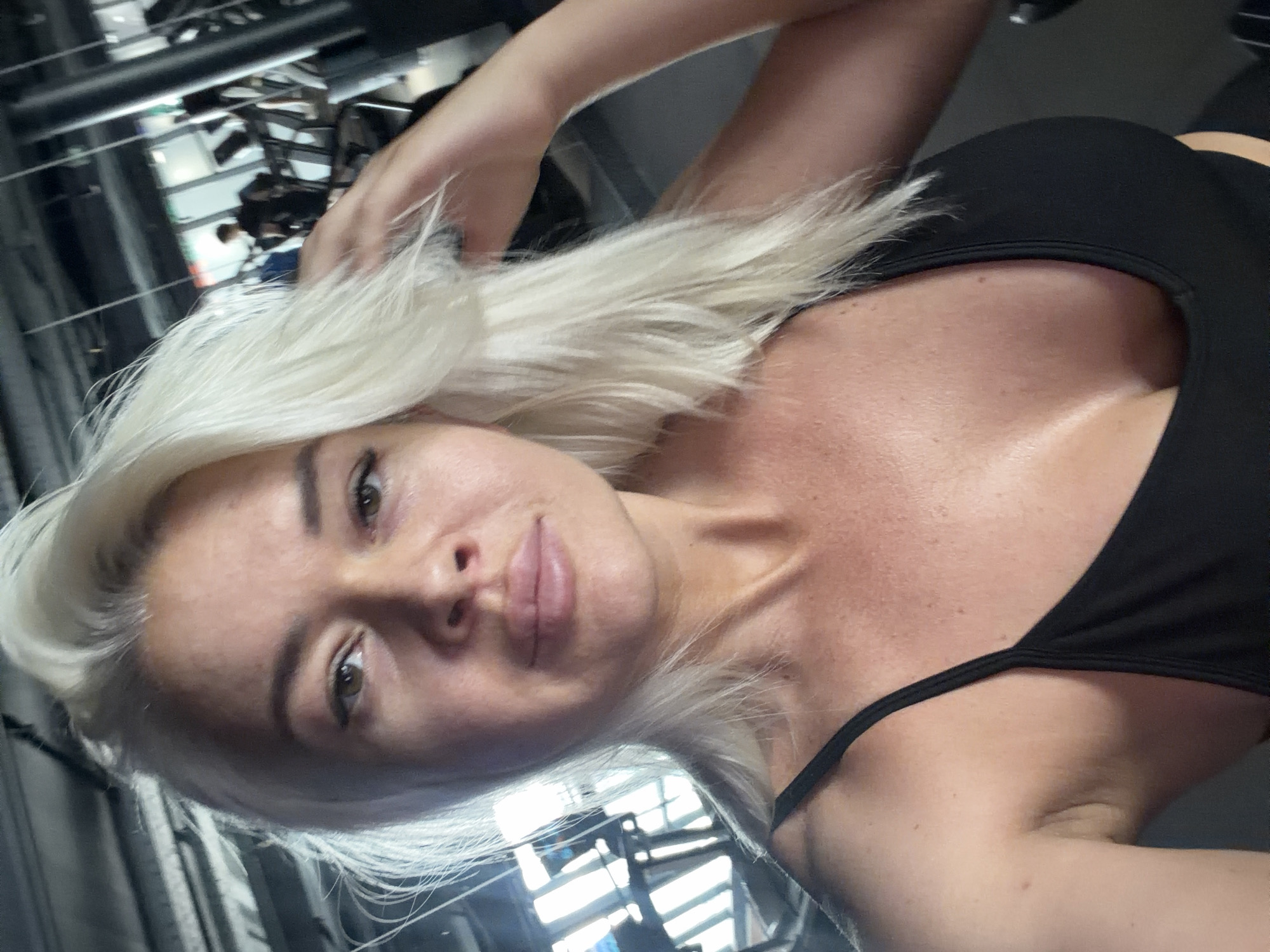 A woman with blonde hair and light skin is taking a selfie in a gym. She is wearing a black tank top and her hand is in her hair. In the background, there are gym machines and other people exercising. The lighting is dim.