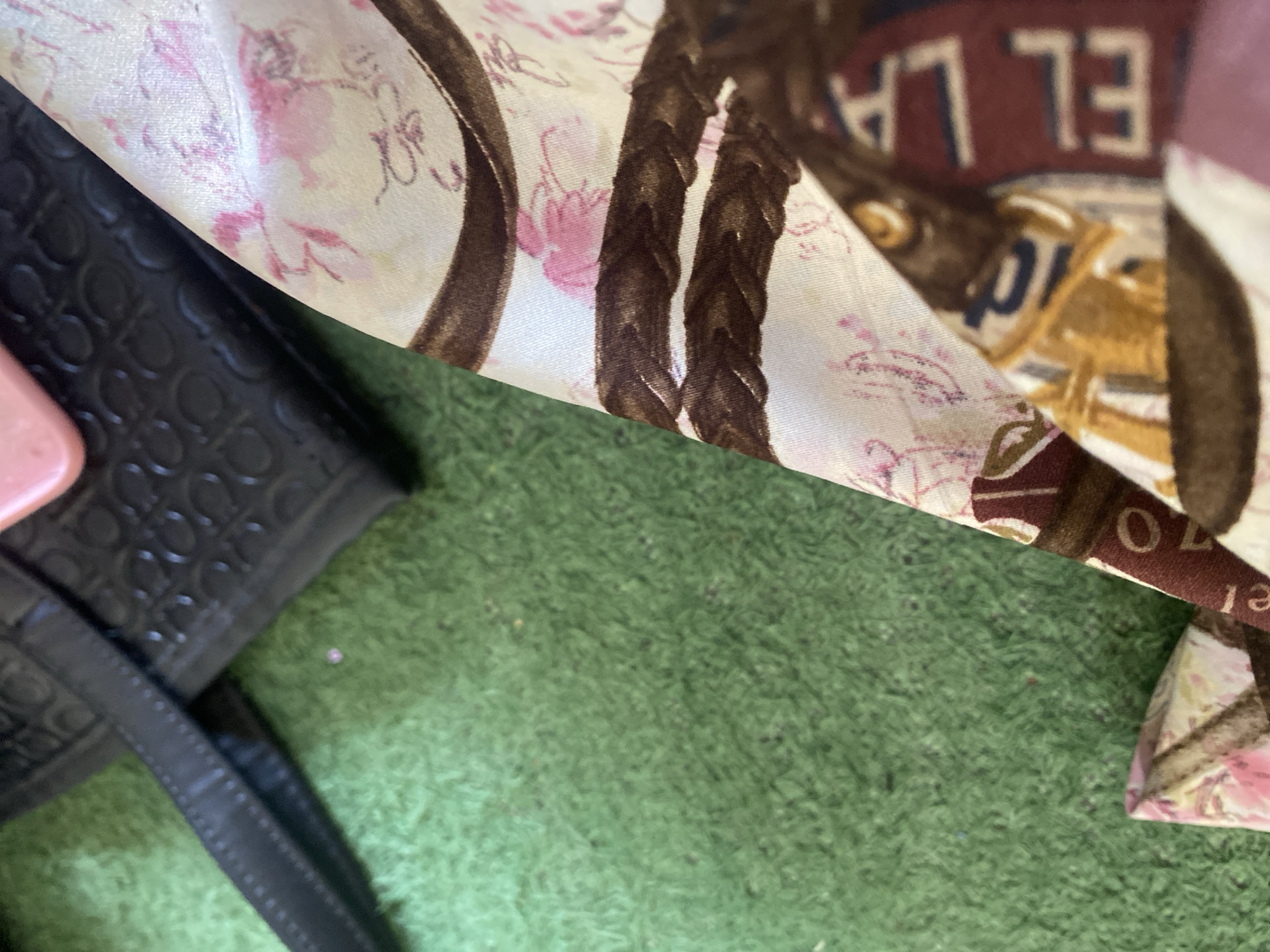 The image depicts a close-up of a patterned fabric, possibly a scarf or a part of a garment, laid on a green, carpet-like surface. The fabric has a light-colored background with floral motifs in pink and brown rope-like patterns. A black, textured material, possibly a handbag, is partially visible in the upper left corner, along with a pink object, likely a phone case. The scene appears to be indoors, as suggested by the lighting and the presence of artificial surfaces. There are no people or discernible activities taking place. The location is not specified within the image.