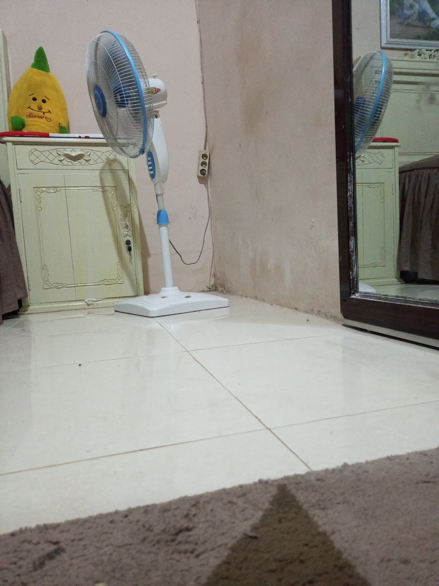 The image depicts a bedroom scene in Lhokseumawe, Indonesia. In the foreground, a plush brown rug lies on a tiled floor. A white pedestal fan with blue accents stands to the right of a cream-colored cabinet, which is adorned with ornate carvings. On top of the cabinet, a yellow plush toy resembling a lemon with a green pointed hat and a smiling face sits to the left of the fan. A power outlet with three sockets is visible on the wall behind the fan. To the far right, a large mirror reflects the room, showing the fan and part of the cabinet. The wall behind the objects appears to be a neutral beige color.