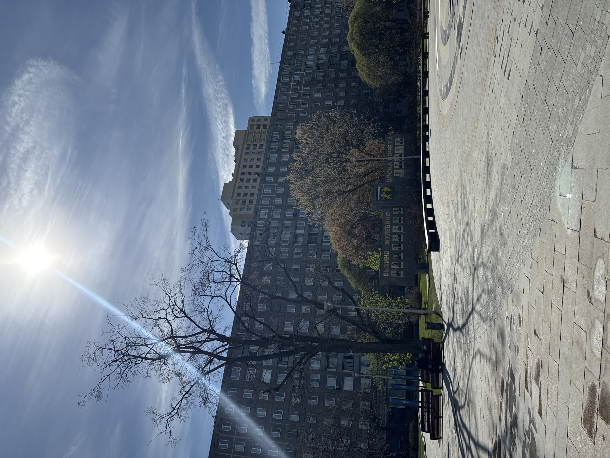 The image depicts an urban scene in Moscow, Russia, characterized by a mix of residential buildings and public spaces. The architectural style suggests the Soviet era with tall, grey apartment blocks dominating the backdrop. A park or plaza is visible in the foreground, featuring benches, trees (some bare-branched, indicating early spring or late autumn), and paved walkways. A notable element is a signboard labeled "ВЫХИНО - ЖУЛЕБИНО ДОСКА ПОЧЁТА," indicating this is a place of honor or recognition for the Vykhino-Zhulebino district. The sky is partly cloudy with the sun shining brightly, casting strong shadows. The overall atmosphere is typical of a residential district within a large city.