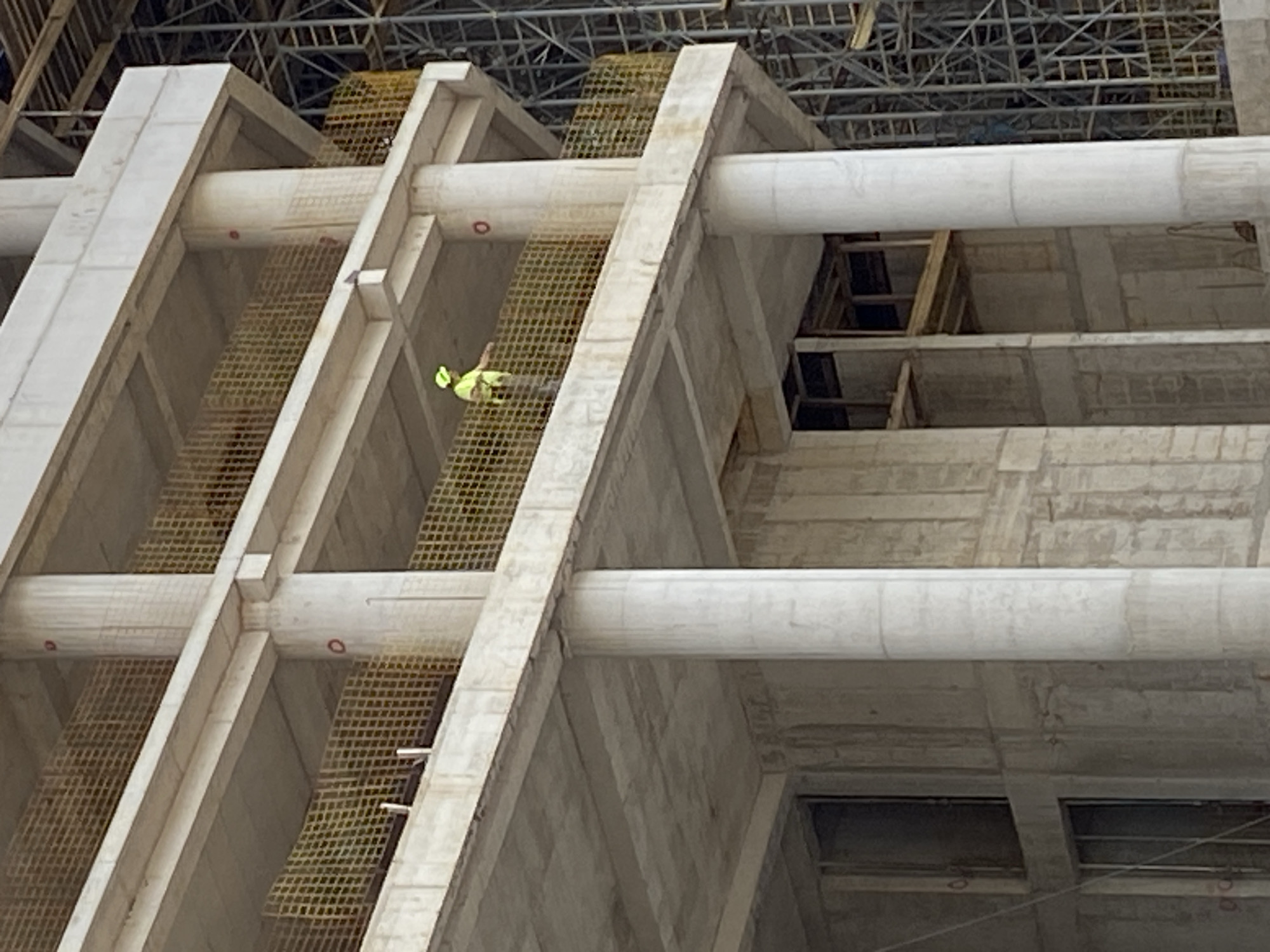 The image depicts the skeleton of a concrete building under construction in Budva, Montenegro. The structure is composed of concrete columns and cylindrical beams. Safety netting is stretched between some of the vertical elements. A construction worker in a fluorescent yellow safety vest is visible within the netting. Above the concrete framework, a metal grid ceiling is partially visible. The overall environment appears to be a construction site.
