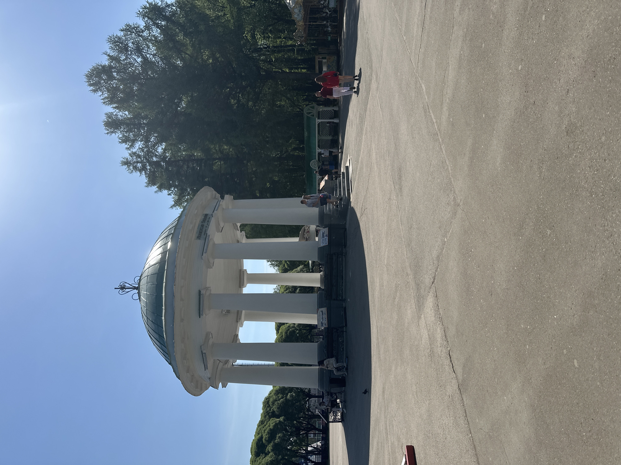 The image depicts a rotunda-like structure with white pillars and a greenish dome, located on a paved surface, likely a square or park area. A couple wearing red shirts is visible in the background. People are walking and seated near the structure. Trees border the scene. The location is identified as Perm, Russia.