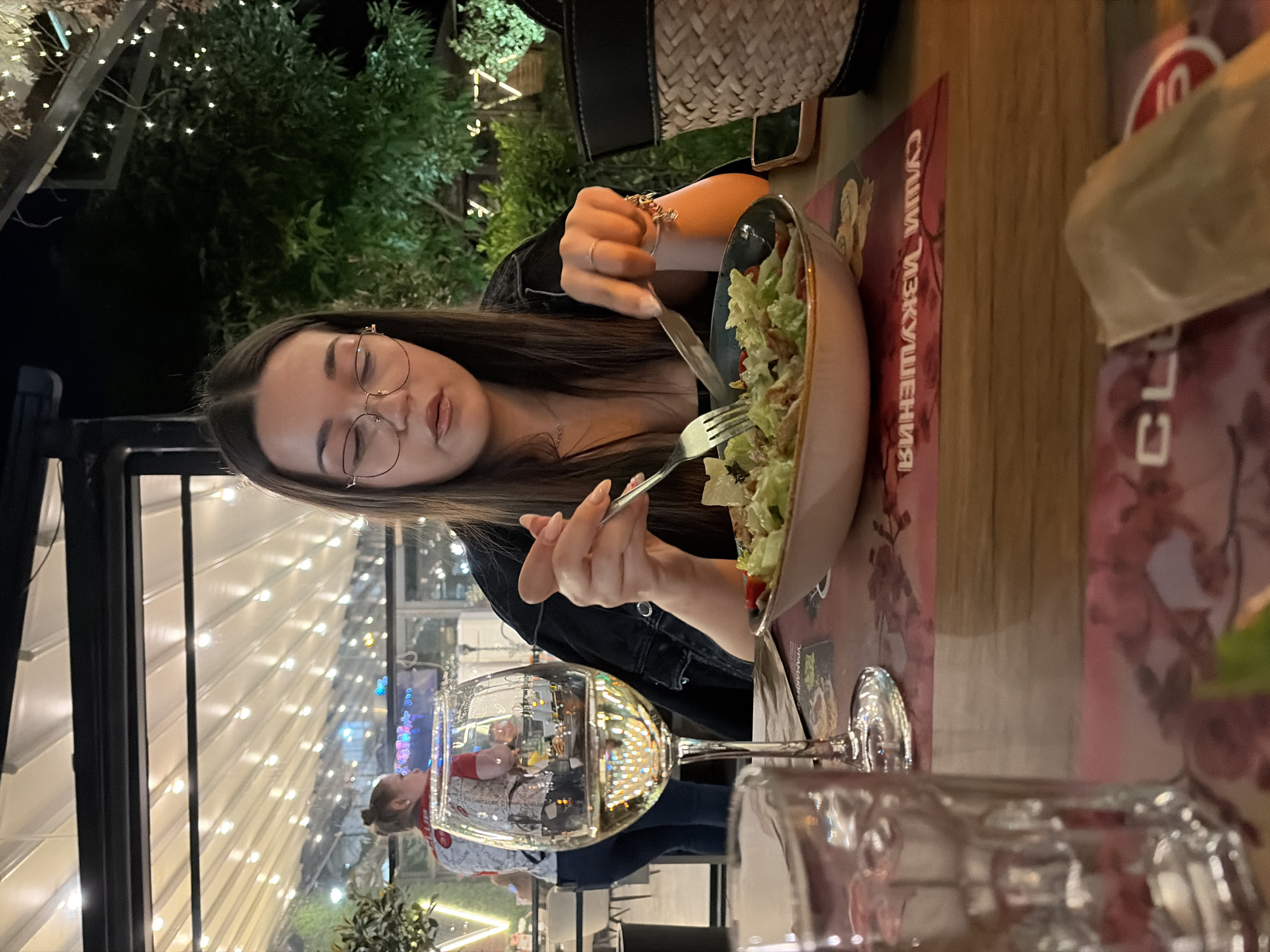 A young woman is dining at a restaurant in Sofia, Bulgaria. She is eating a salad with a fork and knife from a bowl, a glass of wine sits on the table next to the salad. The restaurant has outdoor seating with string lights overhead. A second person is reflected in the glass.
