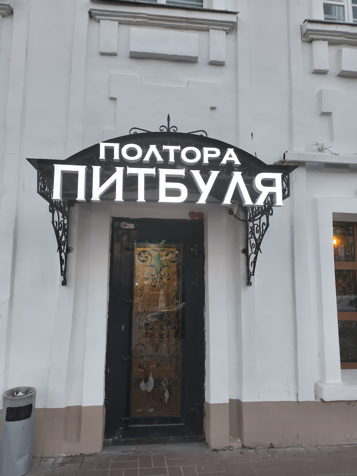 The image shows the facade of a building with a prominent white illuminated sign that reads "ПОЛТОРА ПИТБУЛЯ" (Poltora Pitbulya). The sign is mounted above a dark-colored door with ornate, stained-glass panels featuring depictions of dogs. The door is set within a white stucco wall with architectural detailing. To the left of the door, there is a silver cylindrical trash can. The scene appears to be outdoors, on a paved walkway. The overall impression is of a storefront or establishment entrance. Based on the Cyrillic signage, it is likely located in Russia. The name "Poltora Pitbulya" translates to "One and a Half Pitbulls," which could be the name of a bar, cafe, or shop. Without additional context or landmarks, the specific location in Yaroslavl cannot be definitively determined from the image alone.