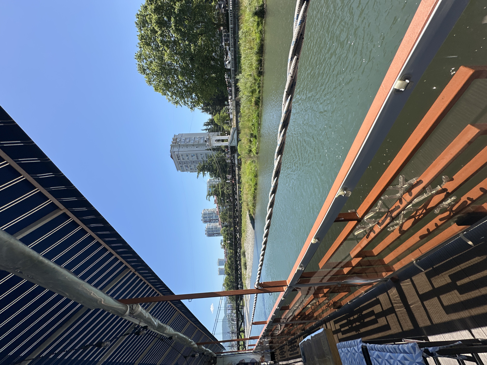 The image depicts a view from an outdoor cafe or restaurant balcony overlooking a river in Sochi, Russia. In the foreground, a wooden deck with tables and chairs is partially visible. The balcony railing is made of wood and glass, with a thick rope acting as a safety barrier. A blue and white striped awning extends over the balcony, casting shadows on the deck.
The river, a murky green color, flows calmly in the middle ground. Across the river, the riverbank is covered in lush green grass and trees. In the distance, several multi-story buildings and a bridge are visible, suggesting an urban or semi-urban environment. The sky is clear and blue, indicating a sunny day. The presence of the bridge and buildings suggests a location within the city of Sochi, likely near the Khosta River or a similar urban waterway. No people are clearly identifiable in the image, and no specific actions or interactions are taking place.