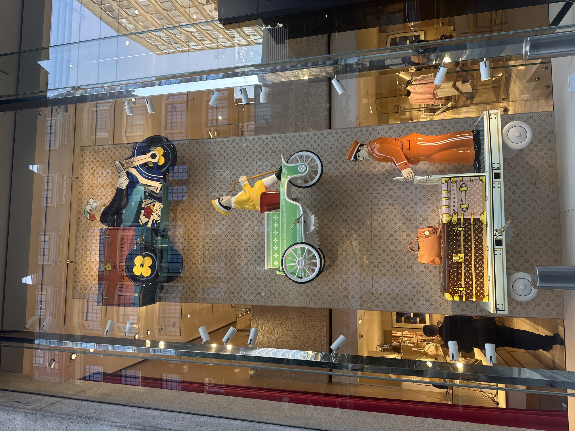This is a full, eye-level shot of a Louis Vuitton store window display in an unknown city. The display features a beige background with the Louis Vuitton monogram pattern. From top to bottom, the display includes a cartoon-like figure riding a blue motorcycle with a red and yellow Louis Vuitton branded truck attached. Below that, a green cart with white wheels is being pulled by a yellow-clad figure. At the bottom, a life-size figure in an orange uniform stands next to two Louis Vuitton trunks, one brown and one pink, on a white platform with wheels. To the left, a man in a dark suit is walking into the store. The window is a large glass pane that reflects the surrounding buildings and sky.