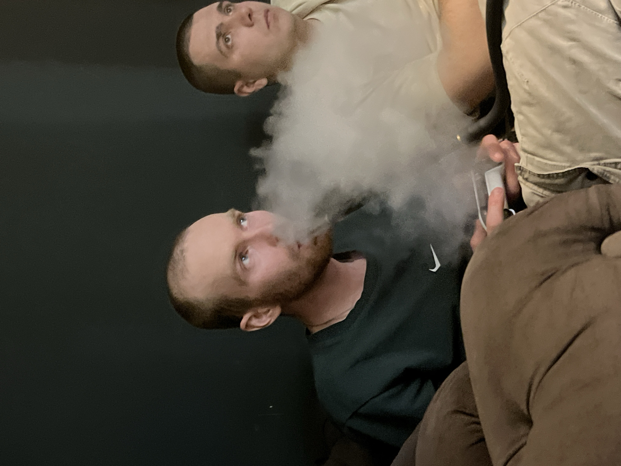 The image captures two men indoors, with one exhaling a large cloud of vapor. The man exhaling is wearing a dark green shirt with a white Nike logo on the shoulder. He has short brown hair and a beard. The second man, located above and slightly to the right of the first, has a buzzcut and is wearing a tan shirt. He holds a white device in his hand. The background is a dark solid color, likely a wall. Other parts of their clothing are visible. The location is Chaikovsky, Russia.
