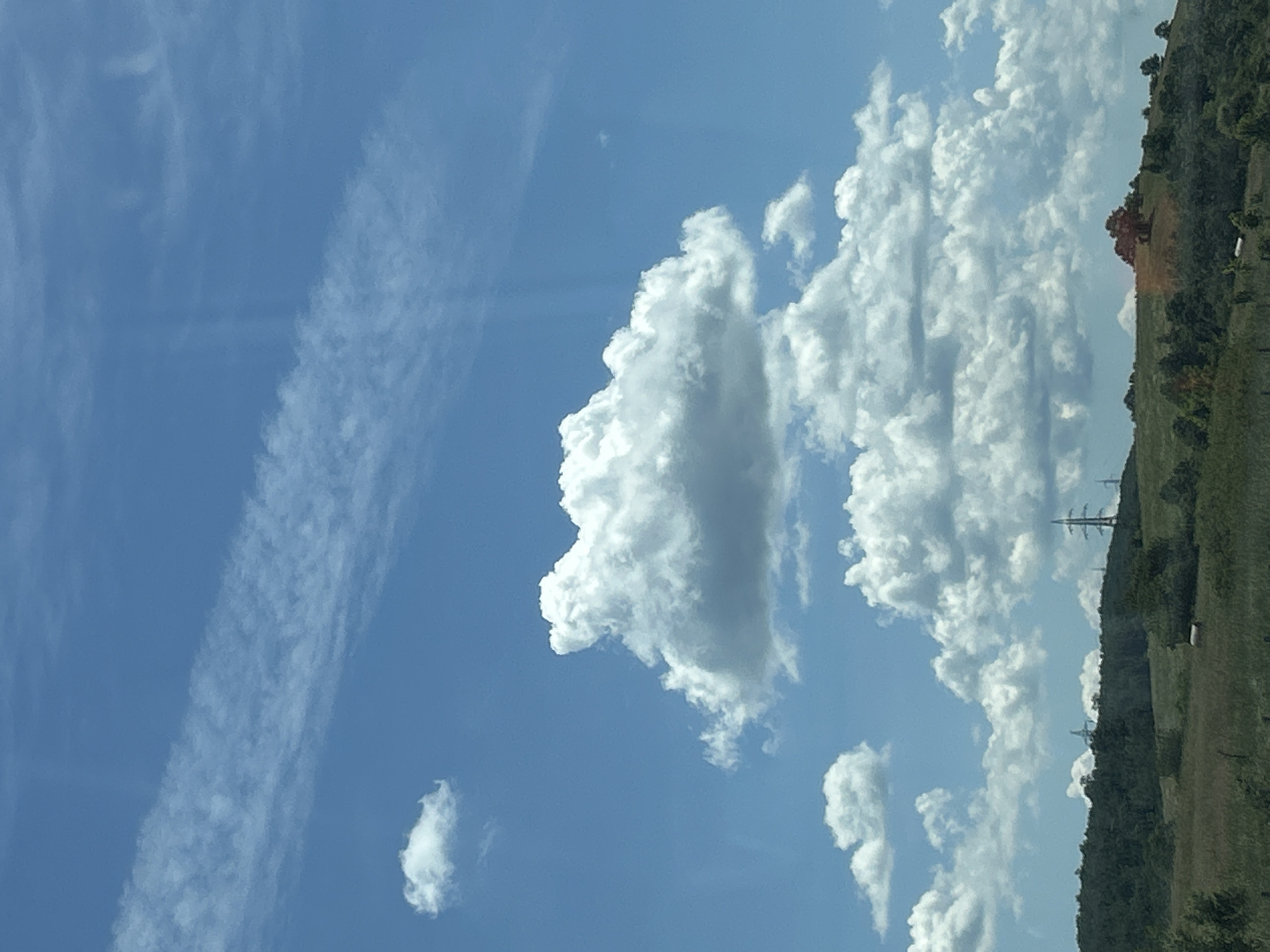 This image depicts a rural landscape under a bright blue sky filled with fluffy white clouds and a thin, wispy band of clouds stretching across the upper portion. The foreground features a grassy field with some scattered bushes and trees. In the midground, rolling green hills are visible, dotted with more trees and some distant structures that appear to be electrical pylons. The overall impression is one of a serene, natural setting, likely a countryside or park area. The location is identified as the city of Rajana, Bulgaria.