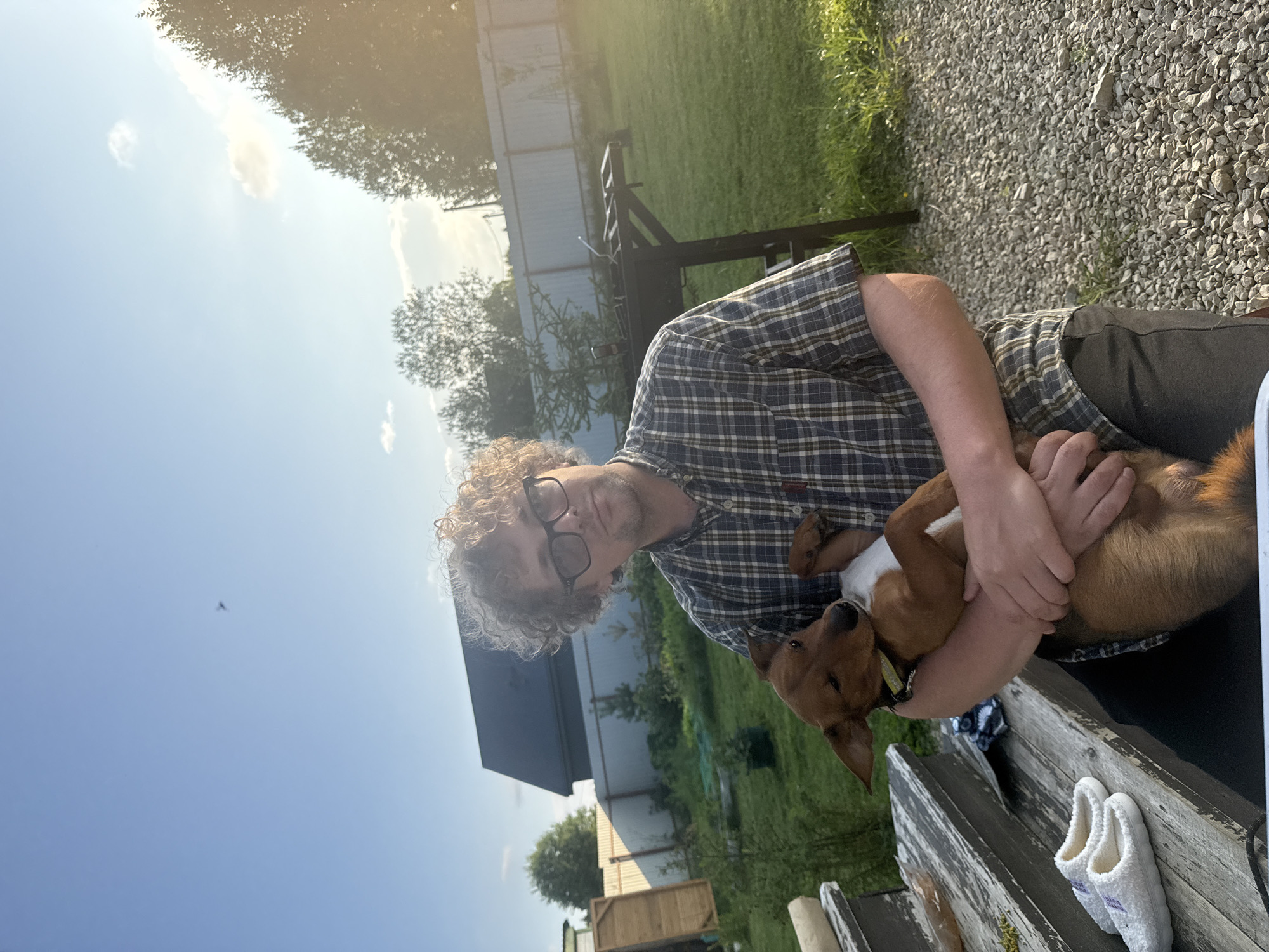 A man with curly hair and glasses, wearing a plaid shirt, holds a small brown dog in his arms. The man is looking at the camera, while the dog is looking away. They are in a backyard with a fence and trees in the background. There are also a pair of slippers on some wooden steps. The sun is setting, casting a warm glow over the scene. The location is the city of Lyady, Russia.