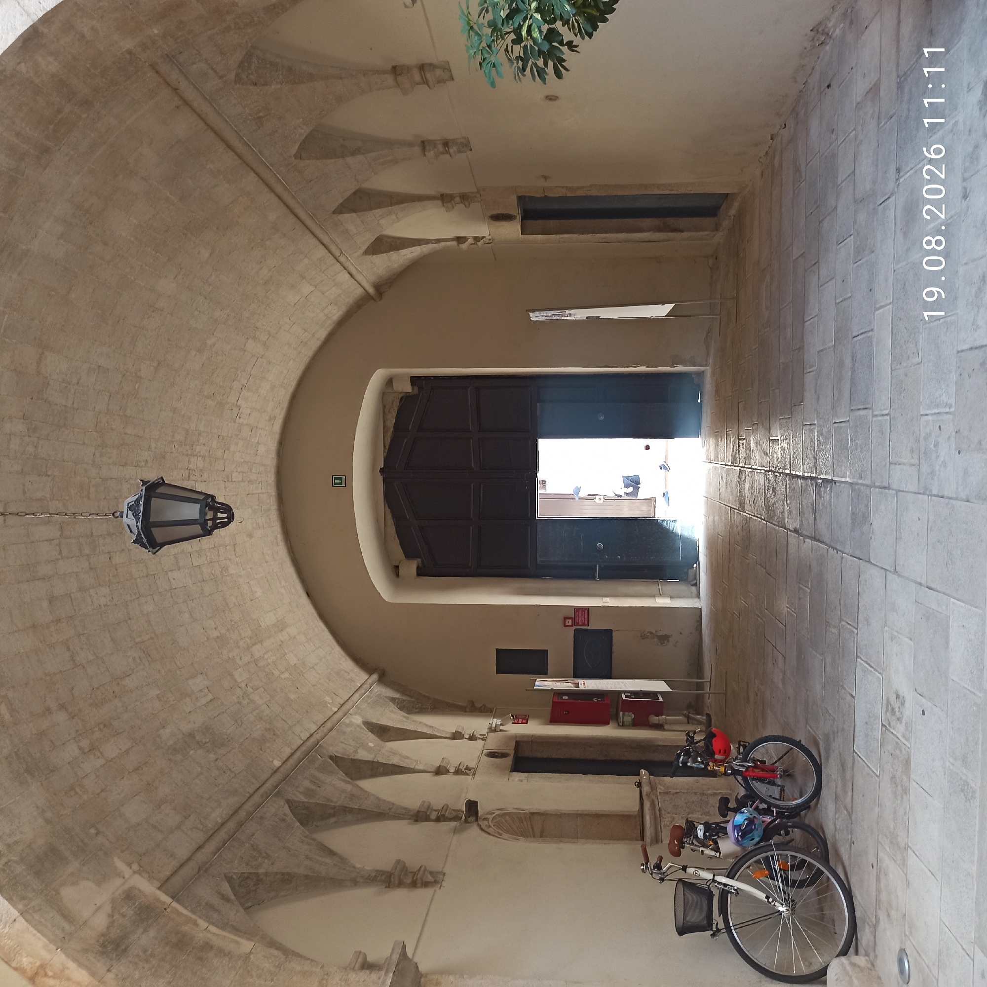 The image depicts a covered walkway with a stone arched ceiling, characteristic of the historic center of Ruvo di Puglia, Italy. Two bicycles are parked on the left side of the walkway, leaning against a wall. The larger bicycle, in white, has a basket on the front and a brown saddle. The smaller bicycle, in red, is positioned behind it and has a red helmet attached to the handlebars. The walkway is paved with rectangular stone tiles, some of which appear wet or reflective due to light. Further down the walkway, a large set of dark double doors is partially open, revealing a brightly lit interior space where a person, wearing a blue cap and white clothing, is visible standing. On the wall to the left of the open doors, there are several signs and a red fire extinguisher. A traditional lantern hangs from the arched ceiling in the upper center of the frame. The overall scene conveys a sense of historical architecture and daily life within a pedestrian area. The timestamp "19.08.2026 11:11" is overlaid on the bottom right of the image.