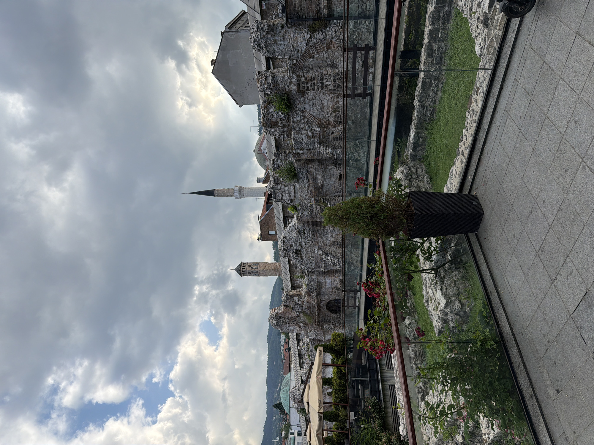 The image depicts a cloudy day in Sarajevo, Bosnia and Herzegovina, likely in the historic Baščaršija district. In the foreground, a tiled patio area is visible, with a low glass railing. Beyond the railing, there are remnants of an ancient stone structure, with arched openings, partially overgrown with vegetation. In the middle ground, a wooden walkway traverses the ruins. Further in the background, several prominent buildings rise above the ruins. To the left, a series of beige umbrellas suggest an outdoor cafe or dining area. To the right of the umbrellas, a tall, brick minaret with a pointed spire pierces the sky, and to its left, a domed building with a green roof is partially visible. Behind these structures, a stone clock tower stands out, and further still, a residential area extends up the hillside. The overall scene conveys a sense of historical layering, with modern amenities integrated into or overlooking ancient ruins within a city setting.