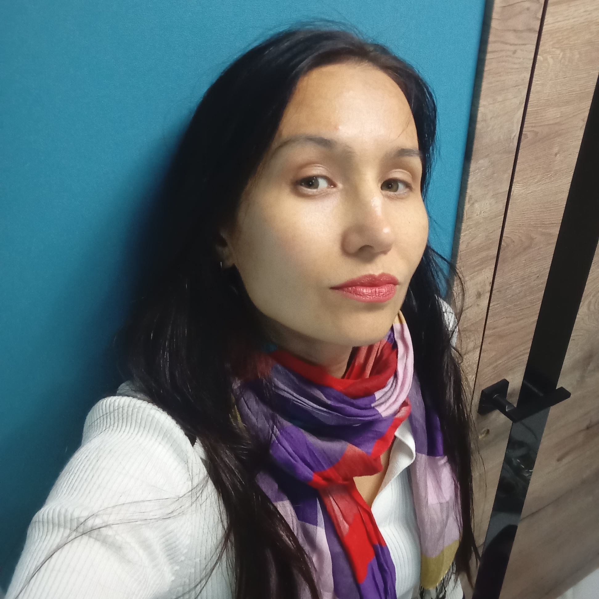 A portrait photograph features a woman indoors in Pervomaysky, Russia, looking directly towards the viewer with a neutral and somewhat thoughtful expression. She has long, dark hair that frames her face, fair skin, and brown eyes, accentuated by red lipstick. She is dressed in a white, ribbed long-sleeved top or sweater, and wears a colorful scarf around her neck. The scarf displays a pattern of red, purple, pink, yellow, and blue geometric shapes or stripes.
The background behind her is split into two distinct sections. On the left, there is a solid teal or blue wall. On the right, a light brown wooden panel, possibly a door or part of a piece of furniture, features a prominent black vertical stripe and a black, angular handle towards the bottom right of the frame. The lighting appears to be soft and even, suggesting an indoor setting with either natural ambient light or artificial illumination. No visible text is present in the image. The woman is posing for the camera.