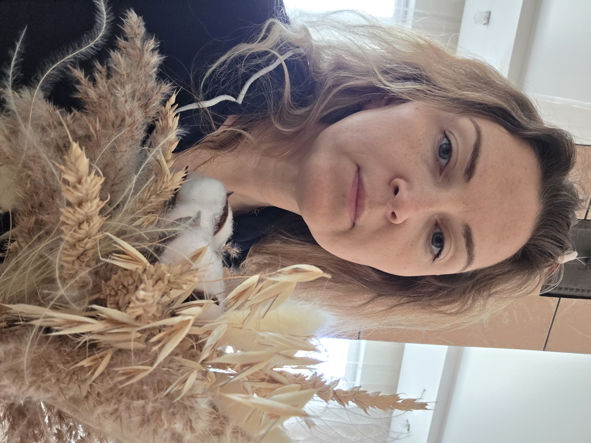 A woman with fair skin and light brown hair is holding a bouquet of dried wheat and cotton. She has blue eyes and a neutral expression. She is wearing a dark-colored top with a white collar. The background is a room with white walls and a reflective surface. The location is not evident and cannot be determined based on the provided image.