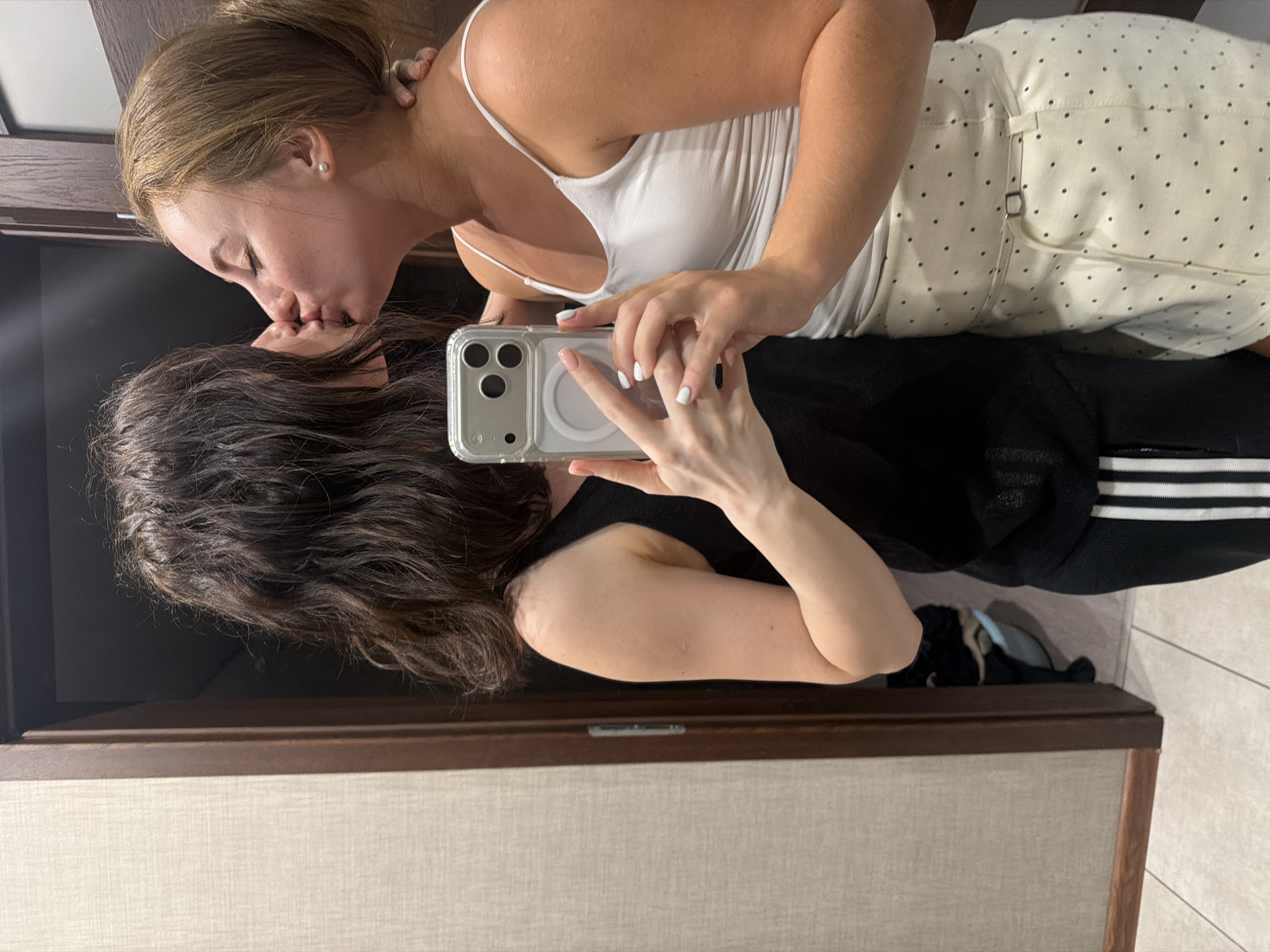 Two women are kissing in front of a mirror. The woman on the left has dark, wavy hair and is wearing a black tank top and black athletic pants with white stripes. The woman on the right has blonde hair pulled back and is wearing a white tank top and a white skirt with black polka dots. She has a pearl earring in her right ear. Both women are holding a smartphone in the foreground. The background appears to be an interior hallway or room with a wooden door frame and textured walls. There is a glimpse of a cat in the background, partially obscured by the woman on the left. The scene is indoors and appears to be a private residence.