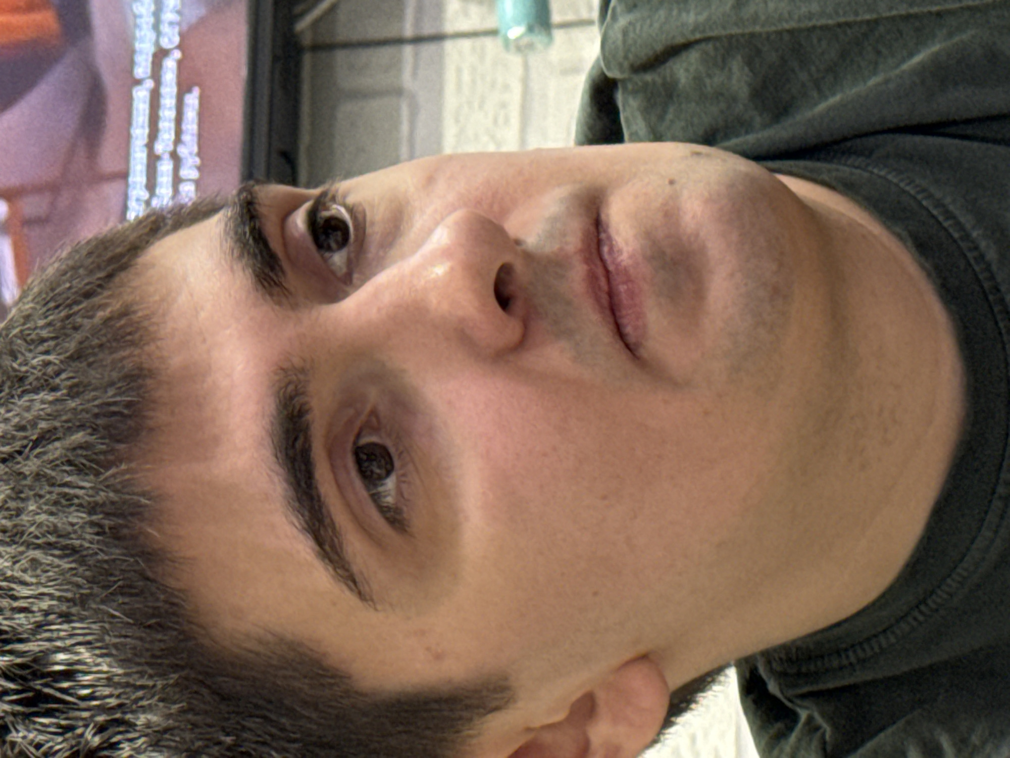 The image shows a close-up of a man's face, angled upwards. He has dark hair, brown eyes, and fair skin. He is wearing a dark-colored shirt. The background is blurred, showing a television screen and other indistinct objects. There is no information in the image to determine its location or connection to Верхнеказымский, Russia.