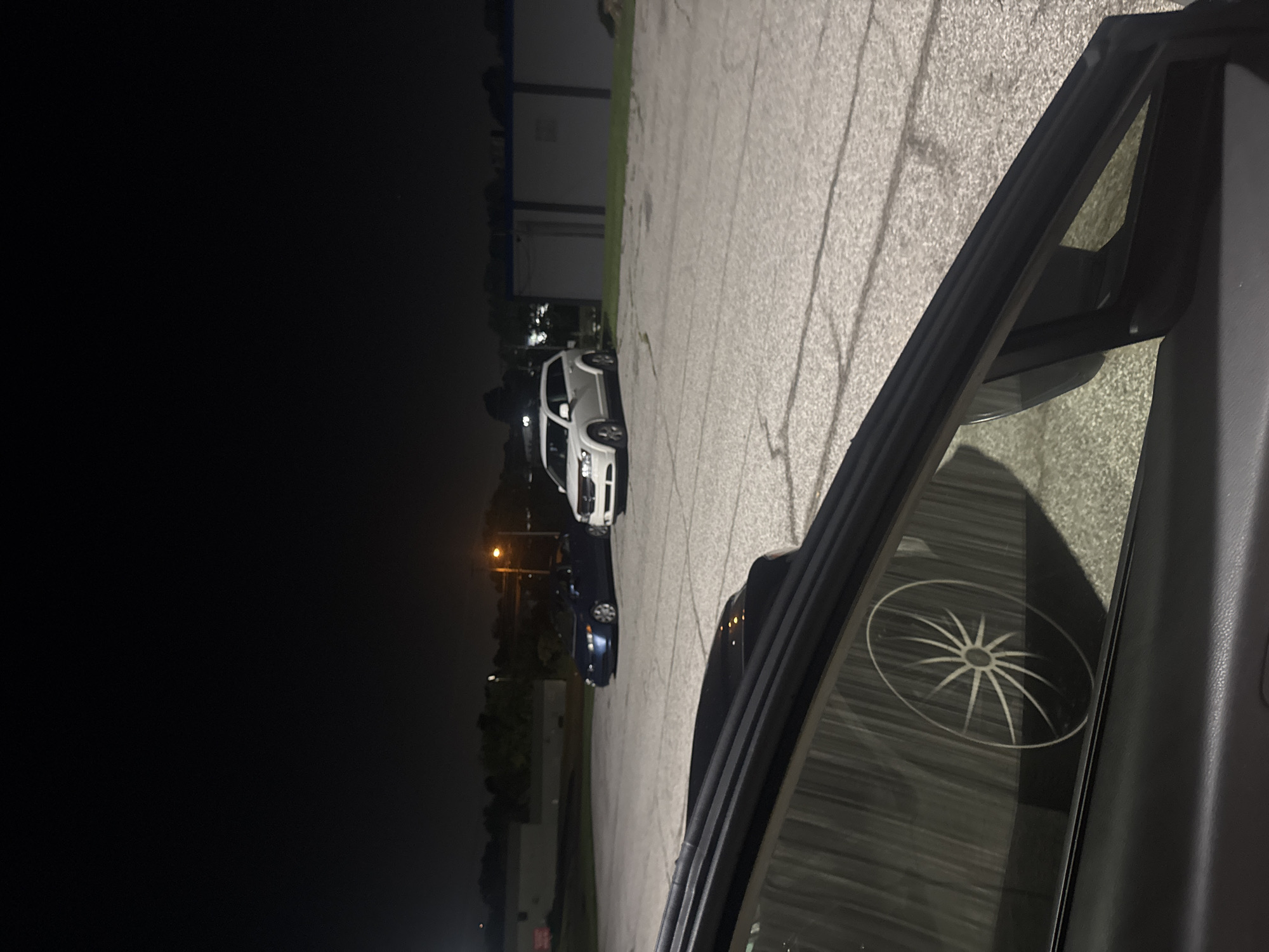 The image depicts a parking lot at night in Jeffersontown, United States of America. A dark-colored sedan is parked on the left, and a white SUV is parked to its right. In the foreground, a portion of a vehicle's interior is visible, including a side mirror and window. The window reflects a graphic design resembling a starburst or a spoked wheel. The parking lot is paved with asphalt and shows signs of wear, with visible cracks and markings. Beyond the parking lot, buildings and trees are dimly illuminated by artificial lights. The sky is dark, indicating nighttime.