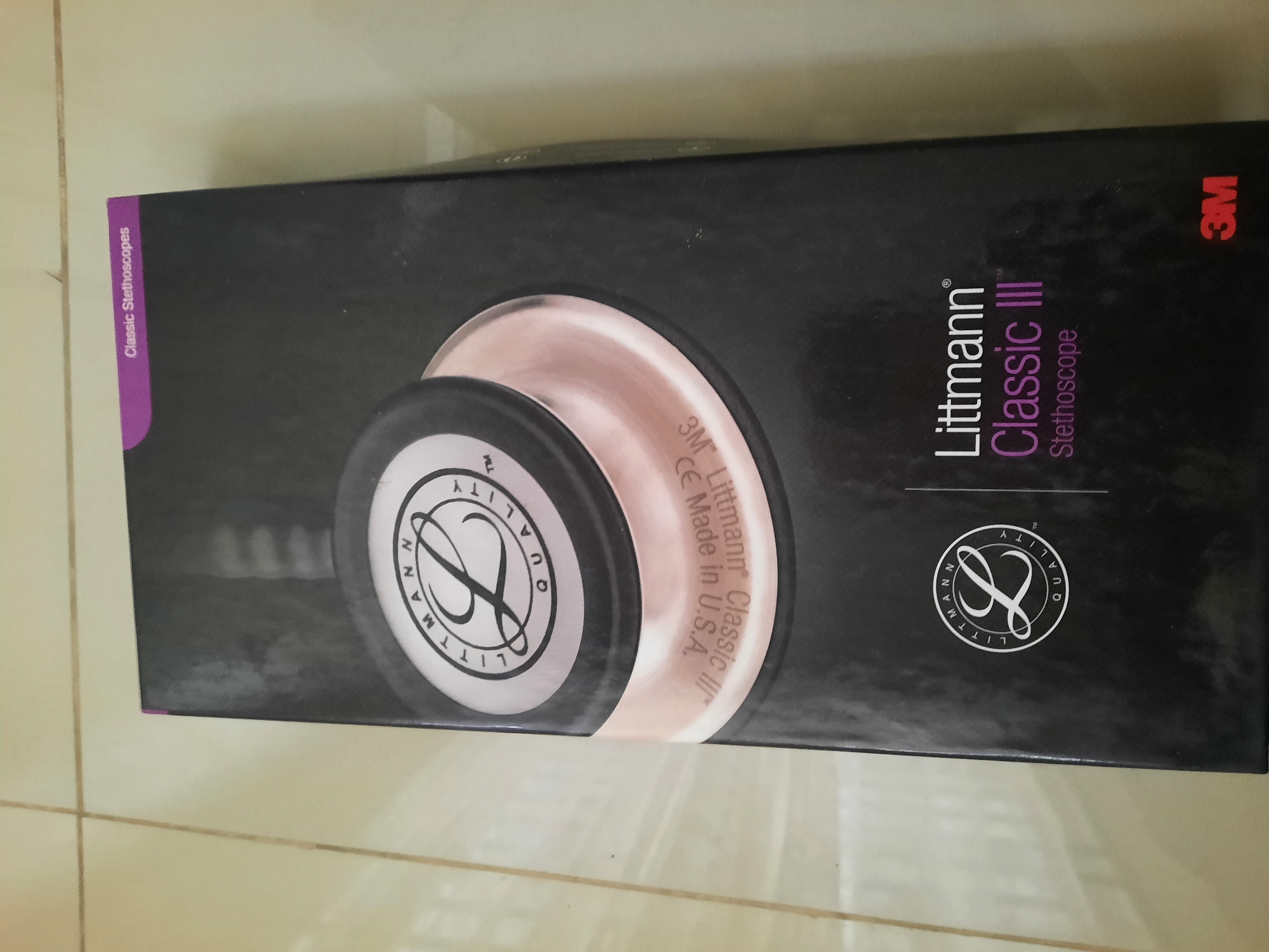 The image shows a closed, black cardboard box for a Littmann Classic III Stethoscope. The box is partially visible, and it appears to be lying flat on a light-colored tiled floor. The top of the box features the Littmann logo, a stylized "L" inside a circle with text that reads "LITTMANN QUALITY TM." Below this, a portion of the stethoscope chestpiece, which is a reflective metallic color, is visible. The box also has text indicating "3M Littmann Classic III Stethoscope." The bottom right corner of the box shows the 3M logo in red. The floor tiles are square and light cream-colored, with visible grout lines. The lighting suggests an indoor environment, possibly a home or a store. There are no people or specific geographical markers to indicate the location in Lhokseumawe, Indonesia.