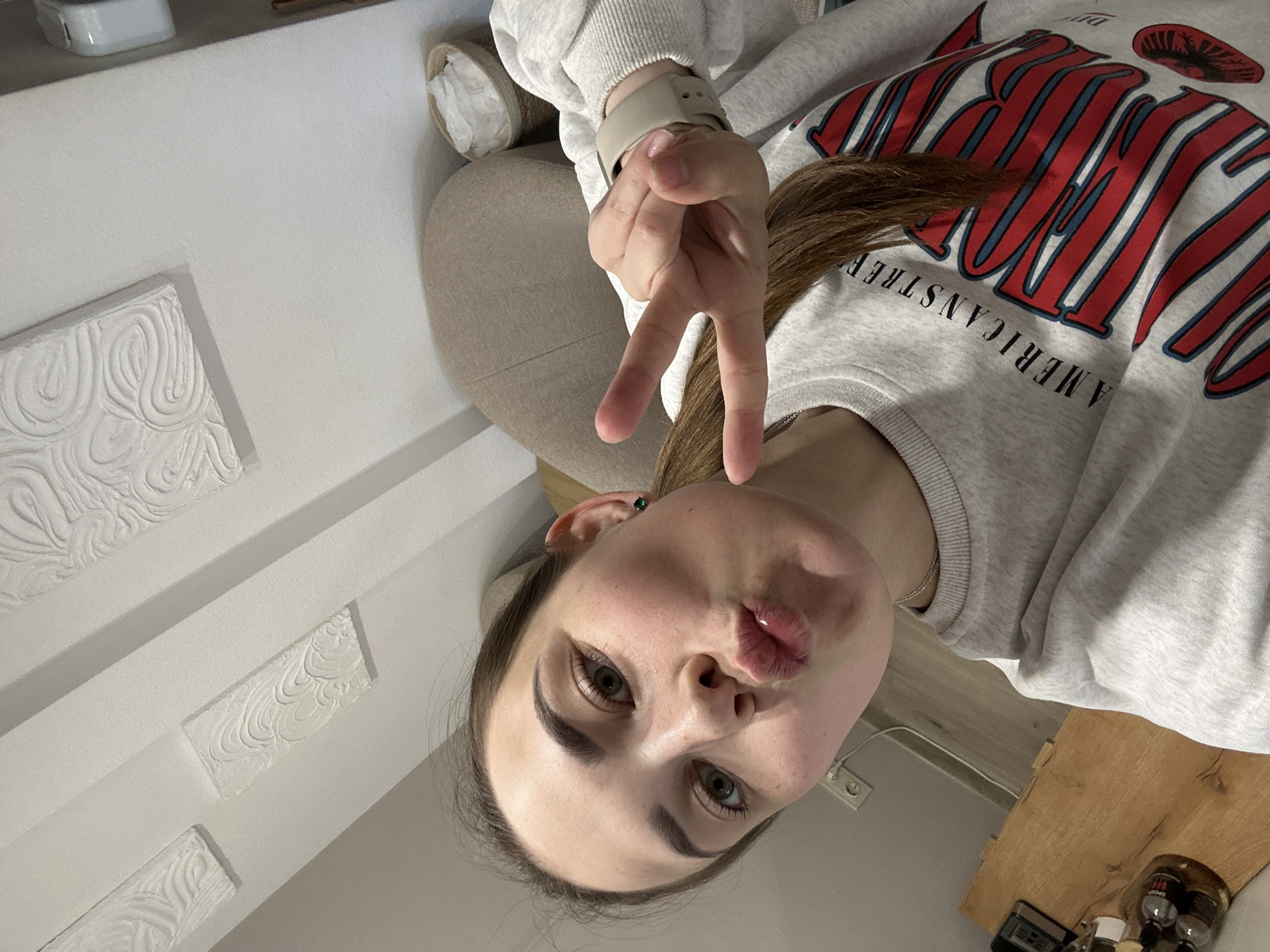 A young woman poses indoors. She wears a gray sweatshirt with the words "AMERICAN STREET" emblazoned on it in red and blue. She is making a "peace" sign with her hand, while simultaneously puckering her lips. The background features textured white walls. Based on the sweatshirt's slogan, and the location in Perm, Russia, it can be inferred the sweatshirt was purchased on American Street (Ulitsa Amerikanskaya). A small table with objects, including several small bottles, is visible in the corner of the frame.