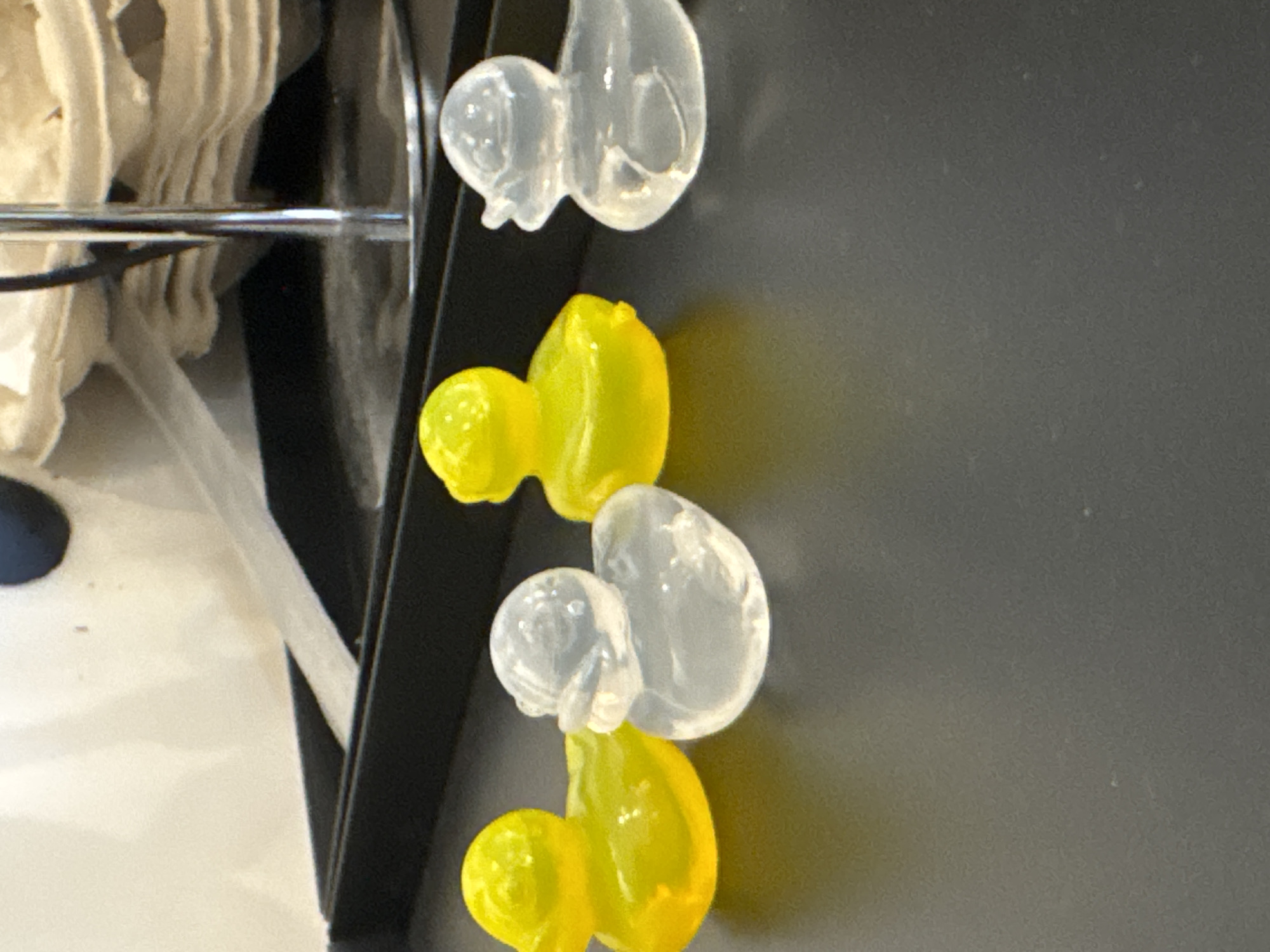 A close-up, eye-level shot shows four small, translucent rubber ducks lined up on a dark, reflective surface. The ducks are arranged from left to right. The leftmost duck is bright yellow, followed by a clear duck, then another bright yellow duck, and finally another clear duck. The background is blurred, with hints of a white, textured surface and a silver pole. There are no people or discernible actions depicted in the image. The scene appears to be indoors, with artificial lighting. The image is a still photograph and does not provide any information about a specific location within the city of Kremenchuk, Ukraine, or any forensic details related to a crime.