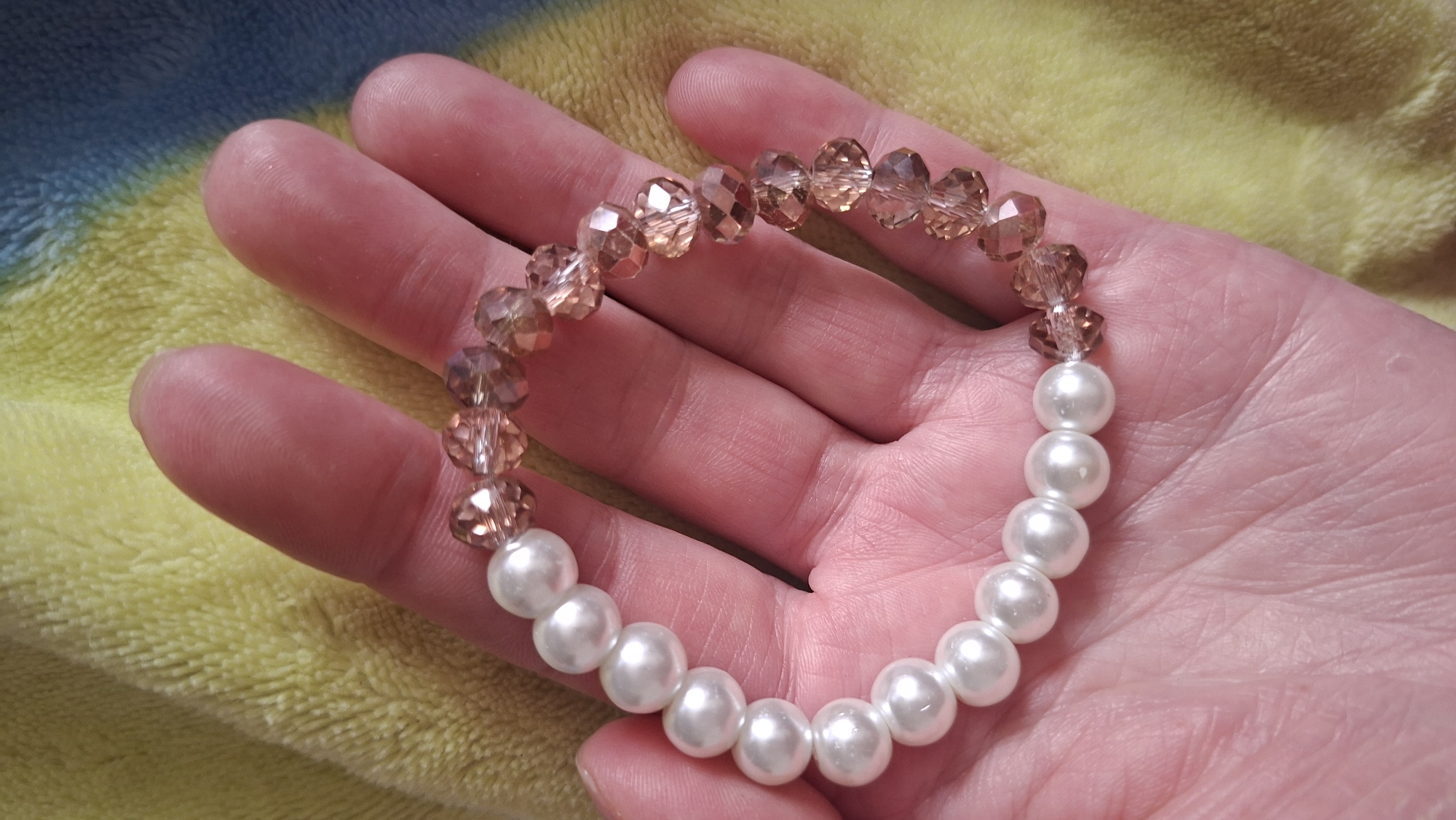 A hand holds a bracelet made of alternating sections of white pearls and faceted brown beads. The background is a blurry, textured surface with a yellow and blue color gradient. The exact location in Забалоцкі сельскі Савет, Belarus cannot be determined from this image.