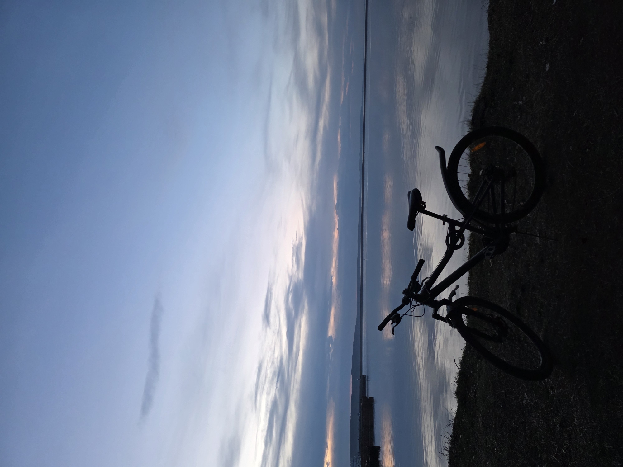 The image shows a bicycle leaning against the edge of a body of water, possibly a lake or reservoir, at twilight. The sky is a mix of blues, grays, and hints of orange and pink, suggesting a sunset or sunrise. The water reflects the colors of the sky. The bike is a dark silhouette, its details obscured by the lack of light. The ground next to the water is covered with grass. It is likely Chaikovsky, Russia, given the prompt.