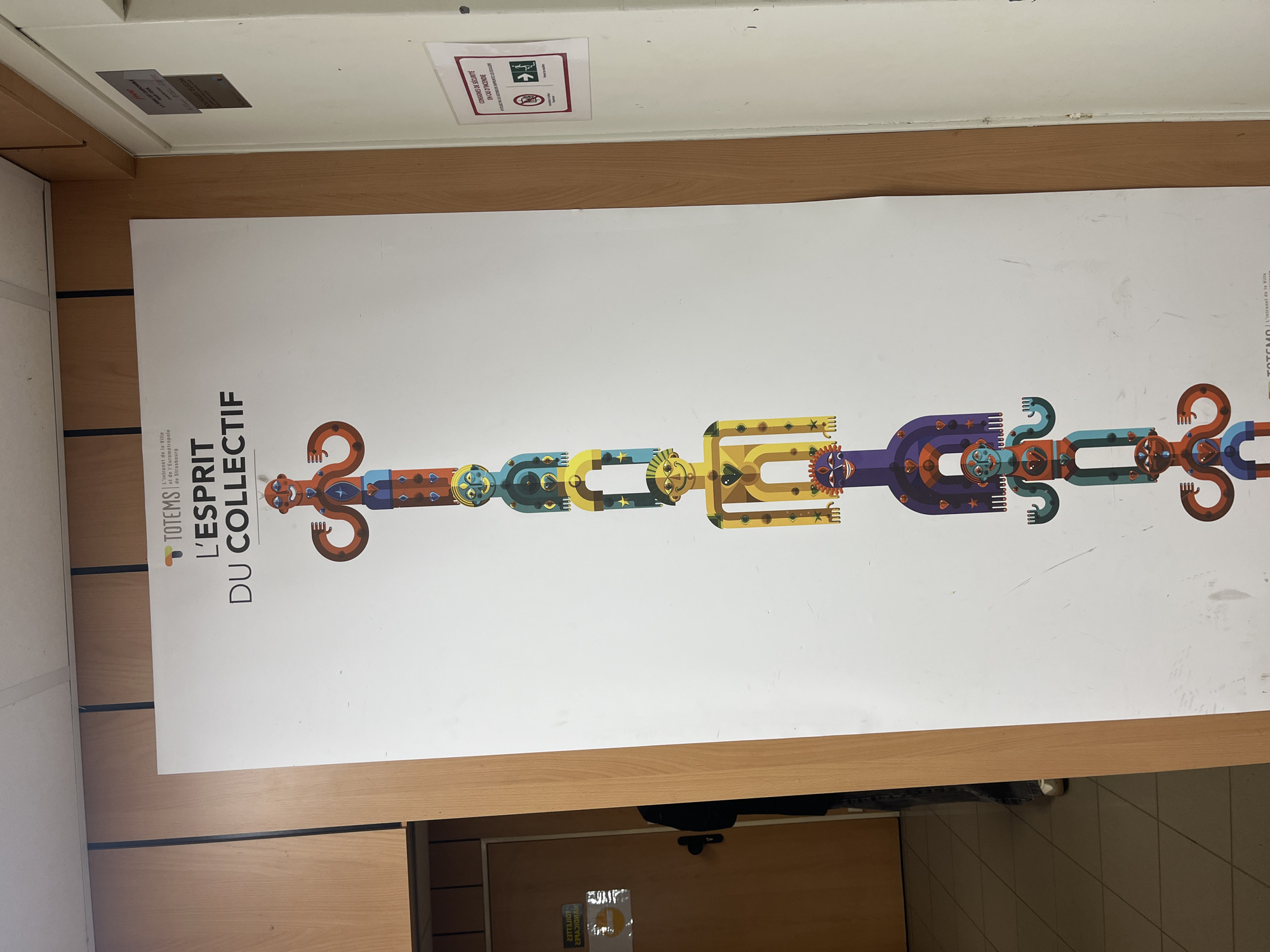 A large, brightly lit indoor scene features a prominent vertical white poster affixed to a wall. The poster's main graphic is a long, horizontal, and highly stylized totem pole composed of abstract, colorful figures or masks. These figures are intricately designed with geometric shapes, vibrant hues of blue, yellow, purple, orange, and brown, and feature elements resembling stars, gears, and flowing lines.
Text on the left side of the poster reads "TOTEMS" with a colorful dot logo, followed by "L'intranet de la Ville et de l'Eurométropole de Strasbourg," identifying its connection to the city administration of Strasbourg, France. Below this, in larger letters, is the phrase "L'ESPRIT DU COLLECTIF" (The Spirit of the Collective), emphasizing community and collaboration. On the far right of the poster, small text includes "TELECOM" and a partially visible copyright or attribution.
Above the poster, the wall transitions to a wooden paneling, topped by a white ceiling. Two small signs are visible on the ceiling: one with "RADIGMAH" and a heart icon, and another displaying "CONSIGNES DE SÉCURITÉ EN CAS D'INCENDIE" (Safety instructions in case of fire), accompanied by pictograms for an exit and no smoking. Below the poster, a dark object, possibly a bag, rests on the floor, next to which a light brown door and a grey tiled floor are visible. The overall impression is that of an informational display within a public or administrative building, likely an office in Strasbourg, France. The scene is well-lit, suggesting daytime.