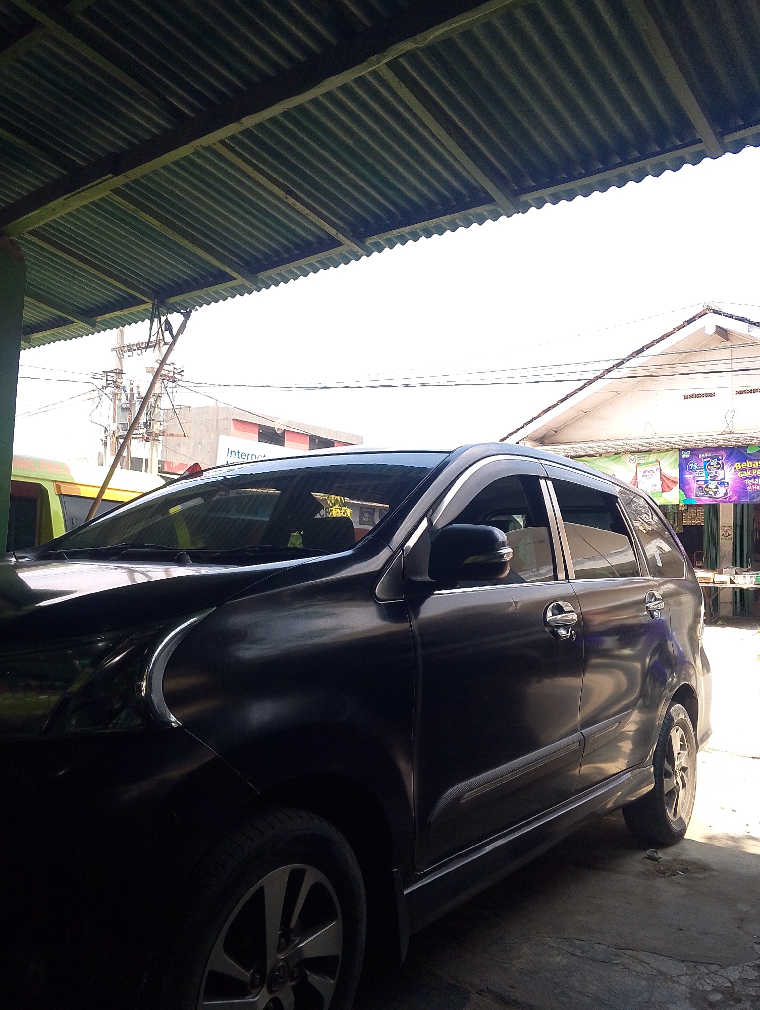 A black, four-door sedan is parked under a corrugated metal awning in a paved outdoor area. The car is positioned in a way that its driver's side is mostly visible. Sunlight streams from the right, illuminating the side of the vehicle and the ground. In the background, a building with a white roof and walls can be seen, along with utility poles and wires. A sign on the building reads "Internet". The overall scene appears to be in a commercial or residential area, likely in the city of Bandar Jaya, Indonesia, given the context. There are no people visible in the image.