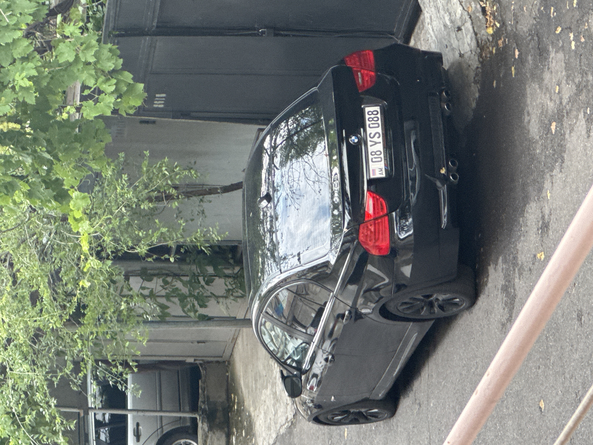 A black BMW sedan is parked on a paved street next to a building with a dark gate. The car's license plate reads "08 YS 088 AM". Overhanging branches with green leaves partially obscure the view of the car and building. A silver car is partially visible behind the BMW.