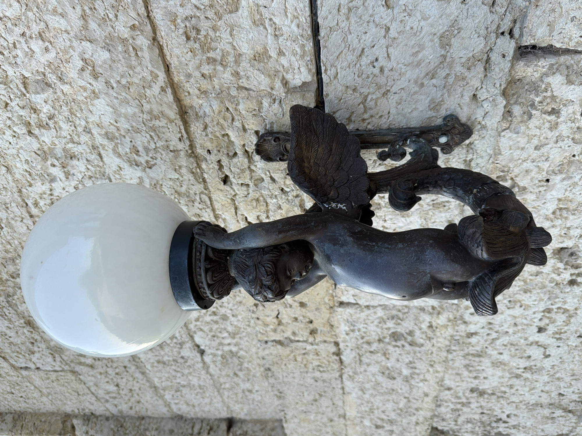 This media file features a close-up, angled shot of a decorative ceiling light fixture. The fixture is made of dark metal and is designed to resemble a cherubic figure with wings. The cherub is depicted in a reclining pose, holding onto a stylized branch that curves around it. The cherub's head is tilted downwards, and its body is smooth and muscular. The wings are detailed with individual feathers. The cherub is supporting a frosted white globe light, which is partially visible on the left side of the frame. The fixture is attached to a stone ceiling, which appears to be aged and weathered, with visible textures and crevices. The lighting suggests it is daytime, and the scene is likely outdoors or in an area with natural light, possibly a courtyard or an arched entryway given the stone construction. There is no visible text in the image.