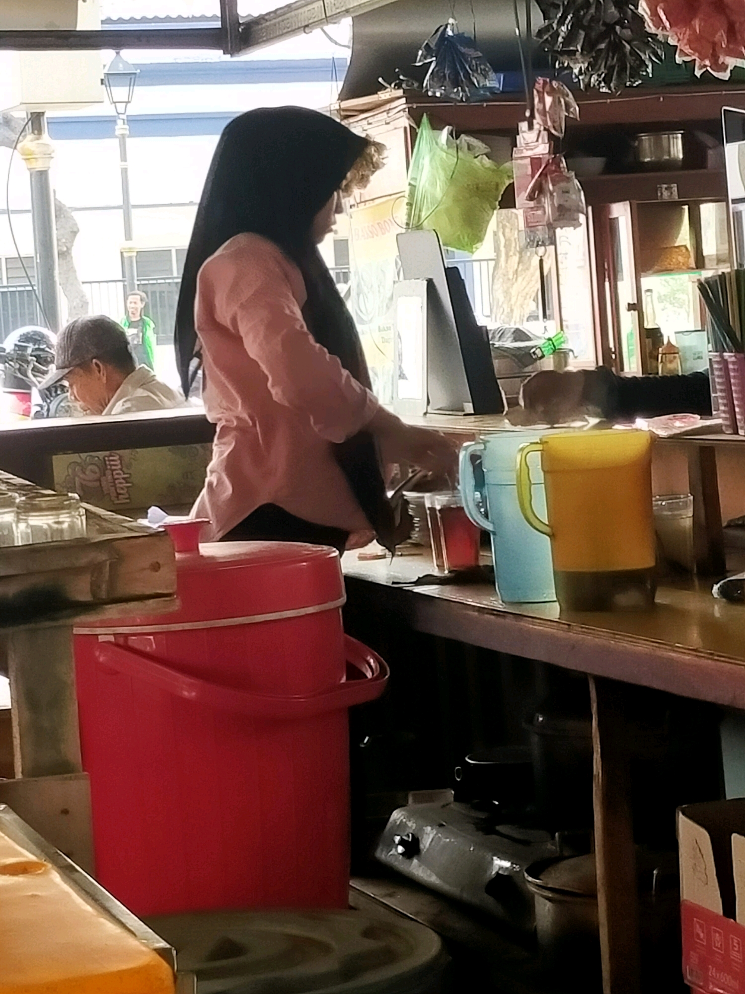 The image depicts an outdoor food stall in Surabaya, Indonesia. A woman wearing a black hijab and a pink long-sleeved shirt stands behind a counter. She appears to be serving or preparing food or drinks. In front of her, on the counter, are several items including a large red plastic bucket, two pitchers (one blue and one yellow), and some cups. Behind the woman, shelves are visible containing various items, including bags and containers. To the left of the counter, a man wearing a cap is seen looking down, and a motorcycle helmet is visible. The background shows an urban street scene with buildings and a lamppost. The overall impression is that of a bustling street food vendor in a typical Indonesian city setting.