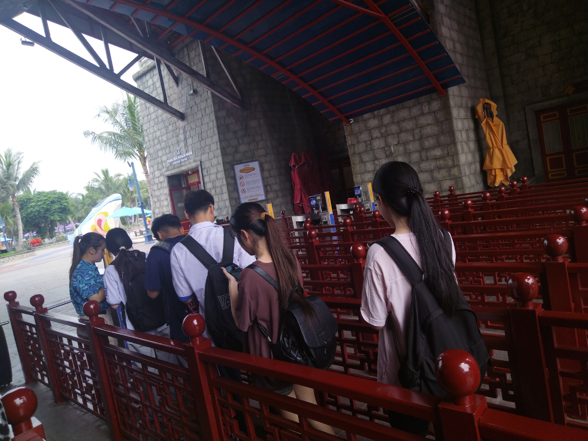 A medium shot shows a line of six people waiting inside an outdoor attraction with a red railing system. The people are mainly young adults, some wearing backpacks. The line stretches toward a stone building with a dark red roof. The building has a sign in a language other than English on its wall, and a yellow robe hangs as if on display. Outside, a glimpse of palm trees and brightly colored structures indicates an amusement park setting. The location is assumed to be in Unknown, Unknown based on the file name.