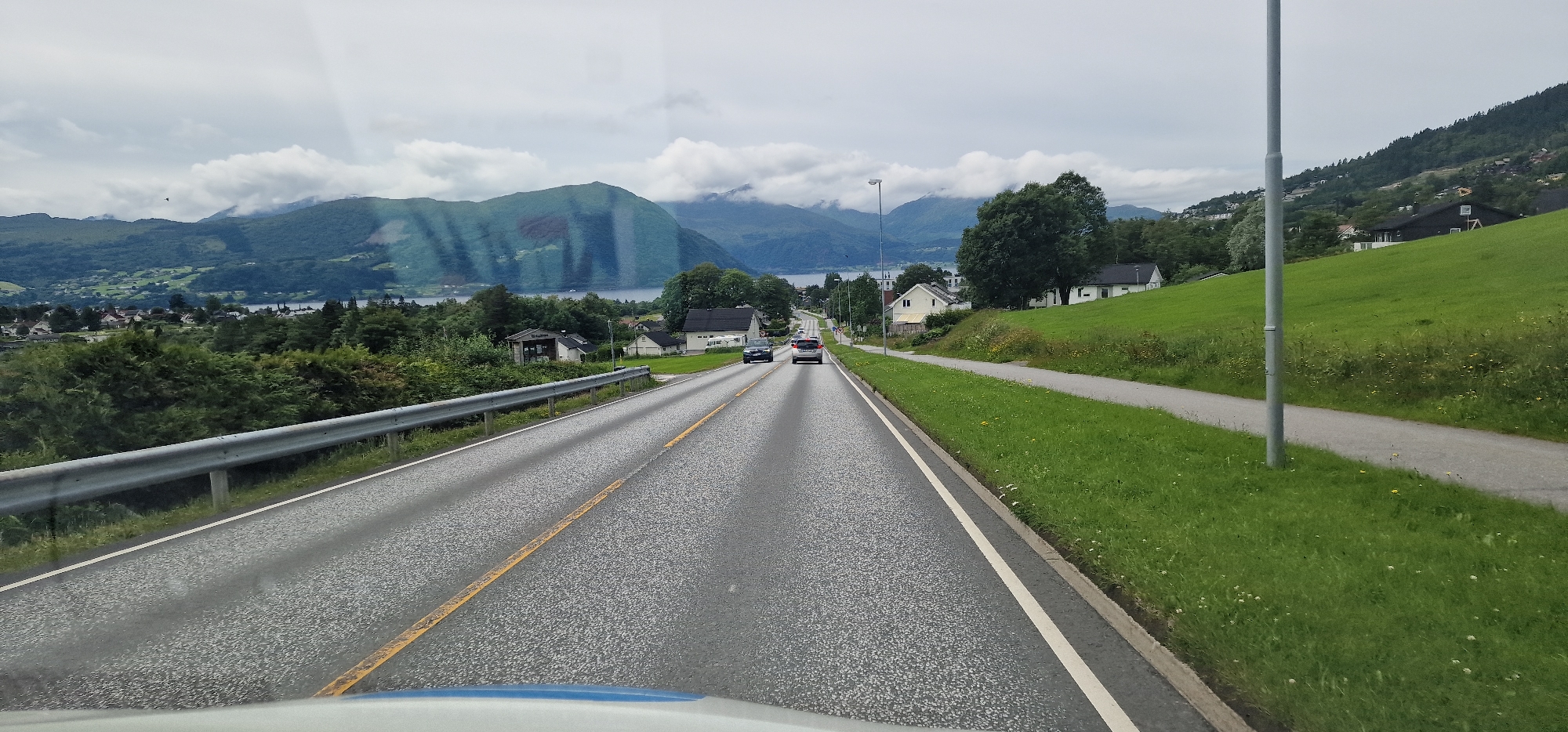 The image shows a road in Volda, Norway, with a body of water and mountains in the background. The road is asphalt and has a yellow line marking the center. There are two cars driving on the road. On the right side of the road, there is a grassy hill with houses and trees. The sky is cloudy.