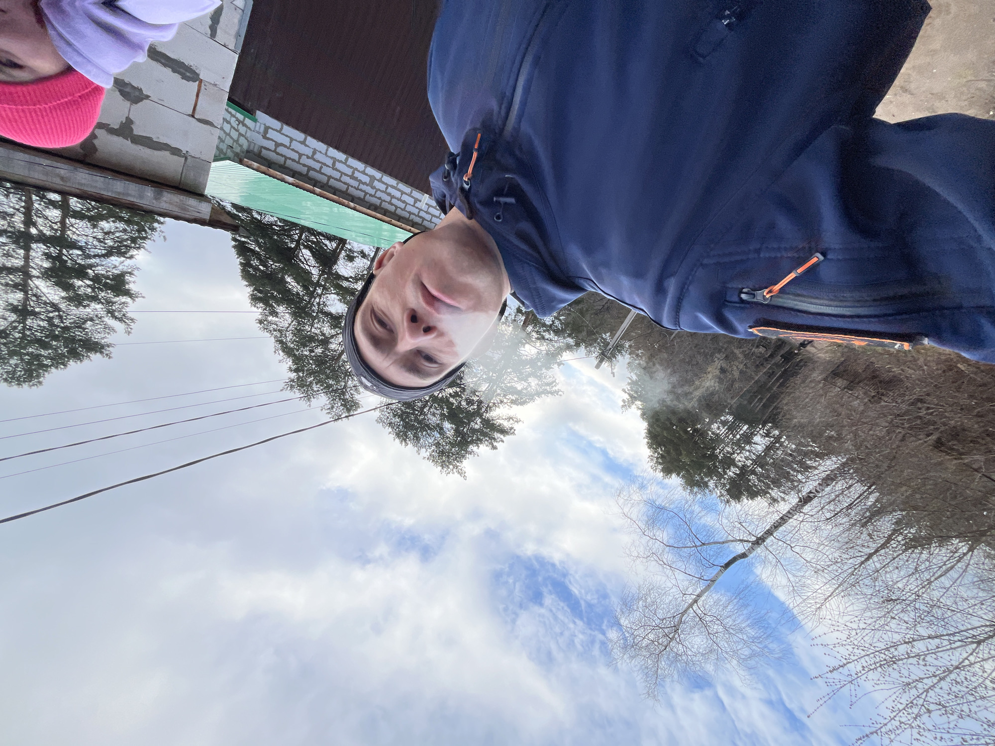 The image is taken from a low angle, looking up. The sky is visible, with blue patches interspersed among white clouds. Power lines run across the sky. A man, wearing a black beanie and a dark blue jacket, appears to be taking a selfie. He is exhaling vapor, possibly from a cigarette or vape. To the left, part of a building is visible, constructed with white blocks and a green roof. Bare trees are visible in the background. Based on the location tag, the scene is in Chaikovsky, Russia. Another person's head is visible in the upper left corner, wearing a pink hat with a white hat.