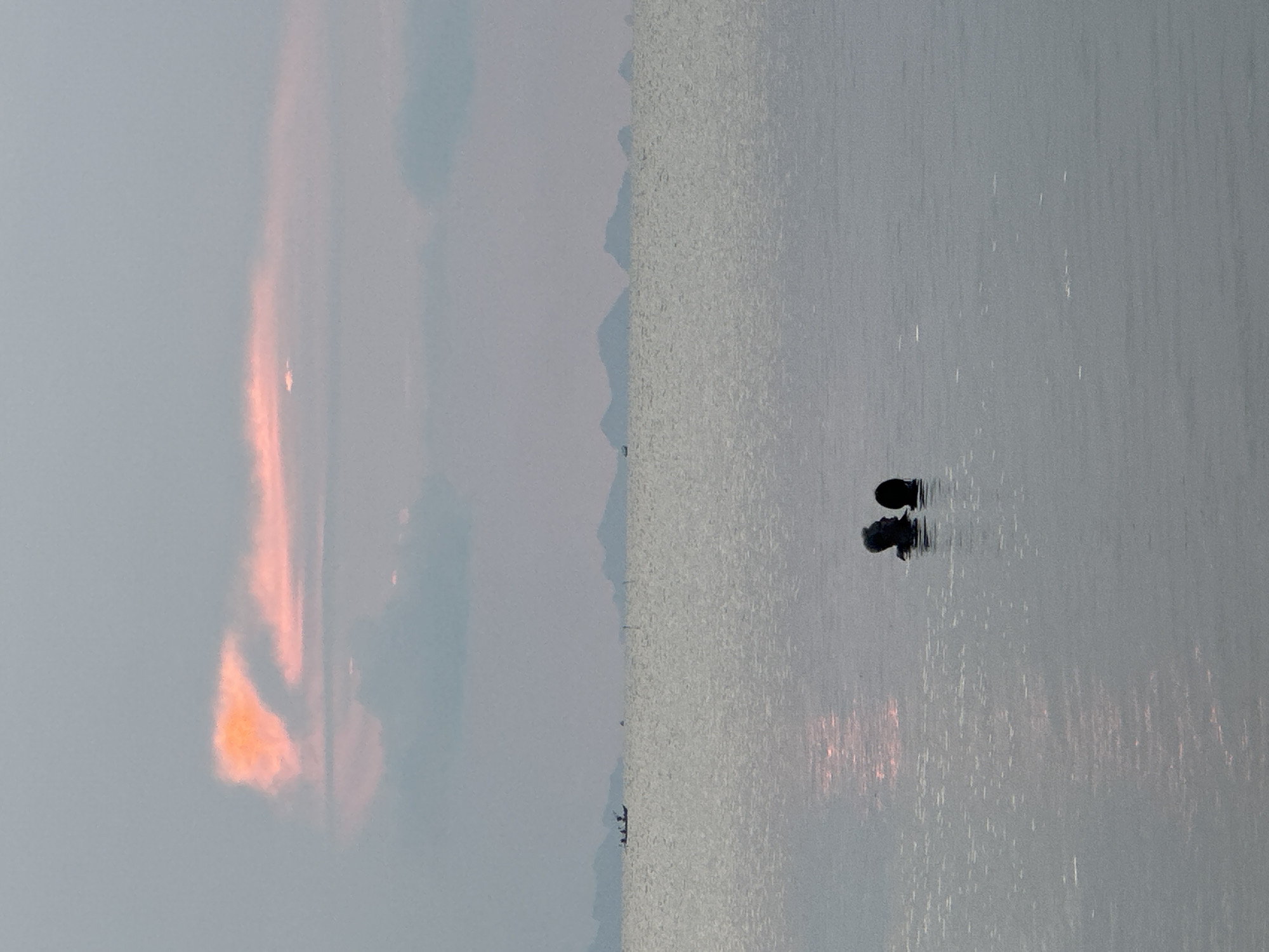 The image depicts a tranquil waterscape at either sunrise or sunset, set in Baan Tai, Thailand. The left side of the frame is dominated by the sky, where a prominent, elongated cloud formation glows with vibrant orange and pink hues, indicating the sun's low position. The remaining sky is a soft, muted blue-gray.
Along the horizon, a faint silhouette of distant mountains or hills stretches across the middle of the image, separating the sky from the expansive body of water on the right. A tiny, dark shape, possibly a small boat or a few individuals in a vessel, is visible near the distant landmass.
The water surface is calm and reflects the warm, colorful light from the sky, creating shimmering streaks and a subtle glow. In the foreground, two dark, silhouetted figures are partially submerged in the water, with only their heads and shoulders visible above the surface. They appear to be people, standing or wading, their forms clearly mirrored in the still water below them. The scene exudes a peaceful and serene atmosphere. No text is visible in the image.