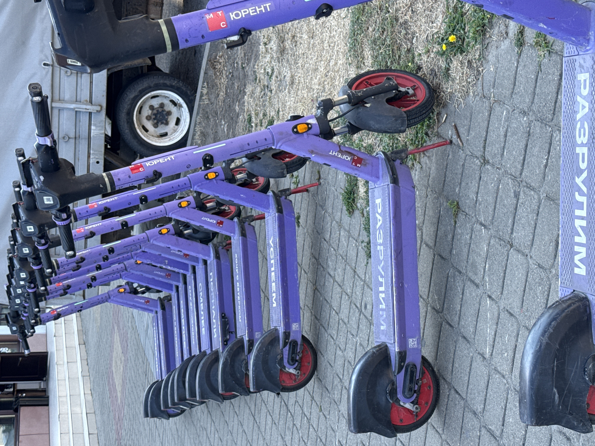 A row of parked, purple electric scooters with black handlebars and red wheel accents are lined up on a paved sidewalk in Anapa, Russia. Some scooters have Cyrillic text printed on their decks, including "ЮRENT" and "РАЗРУЛИМ". In the background, a white truck is parked, and a building with stairs is visible. The ground is a mix of paved stones and dry grass. The lighting suggests it is daytime. There are no people or animals present in the image.