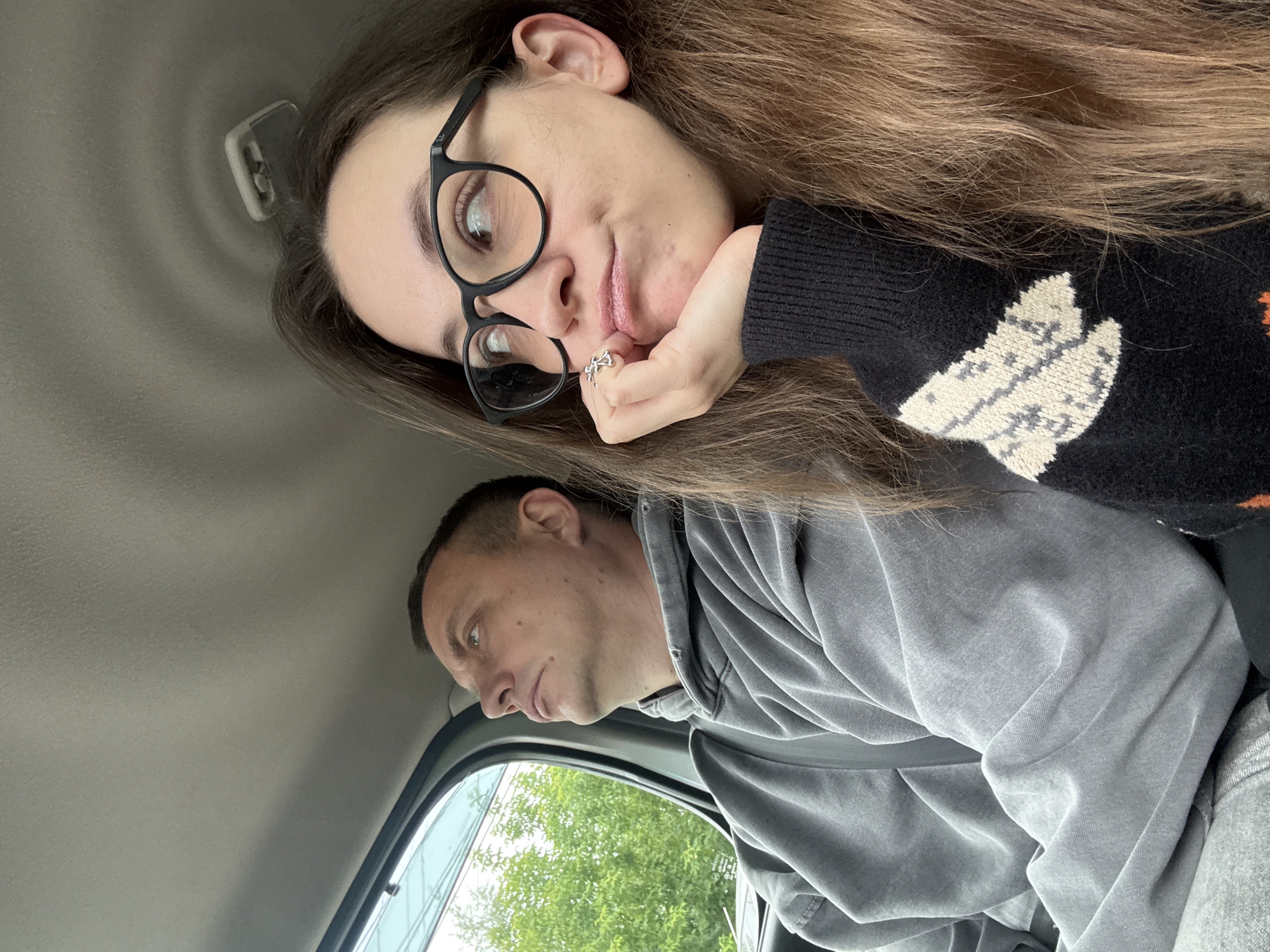 The media file is a selfie taken inside a vehicle, with the camera rotated 90 degrees counter-clockwise. Two people are visible. In the foreground, a woman with long brown hair, wearing glasses and a black sweater with a white pattern, rests her head on her hand. She is looking towards the camera with a slight smile. A ring is visible on her finger. Behind her, a man wearing a grey hoodie is looking out the window to his left. The setting appears to be daytime, with natural light filtering into the car, illuminating the interior and the trees visible outside the window. The overall mood seems casual and relaxed. No visible text is present in the image.