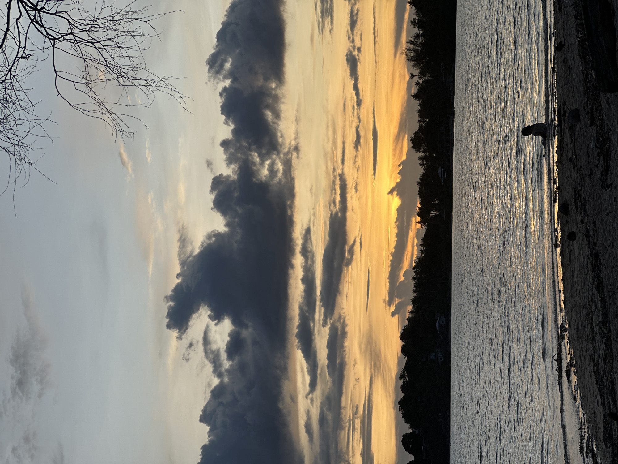 The image depicts a scenic sunset over a body of water. The sky is filled with dramatic clouds, with dark gray formations on the left contrasting with streaks of orange and yellow on the right, indicating the setting sun. Silhouetted trees form a dark line along the horizon. On the right side of the image, the water is calm with gentle ripples reflecting the sunset's colors. A person, partially visible and in silhouette, stands by the water's edge on the sandy shore. The overall mood is serene and peaceful, evoking the beauty of a tropical sunset. The location context suggests this scene is from Koh Phangan, Thailand, which aligns with the lush, silhouetted vegetation. The time of day is clearly sunset, and the weather appears to be clear apart from the clouds. There is no visible text in the image.