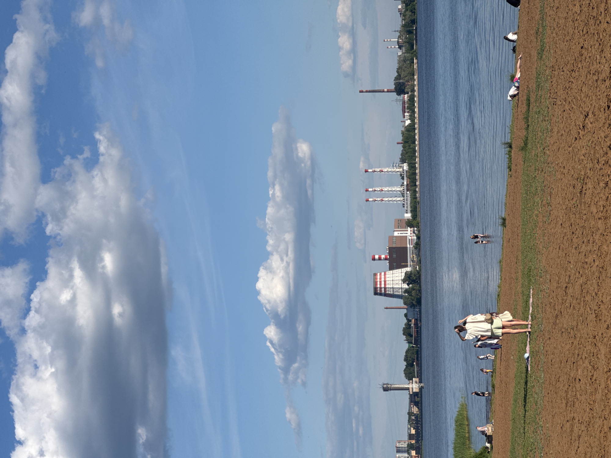 This image depicts a wide-angle scene of a waterfront area in Izhevsk, Russia, under a partially cloudy blue sky. The sky above features various formations of cumulus clouds, ranging from dense grey masses to lighter white wisps.
The immediate foreground consists of a reddish-brown sandy beach with patches of green grass near the water's edge. Several individuals are present. Centrally, a woman, seen from behind, wears a white long-sleeved shirt and light-colored shorts, and carries a light shoulder bag while adjusting her hair. She stands on a light-colored mat. To her immediate right, another person is partially obscured. Further right on the sand, an individual in a white top and pink shorts lies prone, facing the water, next to another person seated upright. To the left, near the water, two people are sitting or leaning on the grassy bank, while several others wade in the shallow water, including one person in dark clothing and another in lighter attire. Two additional individuals are wading further right in the water.
The midground is dominated by a wide, blue-grey body of water, exhibiting subtle ripples across its surface.
The background presents an industrial skyline across the water. On the left, a tall, slender grey water tower with a conical top is prominent. To its right, a large cooling tower with distinct red and white horizontal stripes stands next to brown industrial buildings. Further along the horizon, a cluster of three tall, slender smokestacks, also featuring red and white striped tops, is visible. Another solitary, tall chimney with similar red and white stripes is positioned to the right of the cluster. Numerous other industrial structures, buildings, and a line of green trees are interspersed along the distant bank.