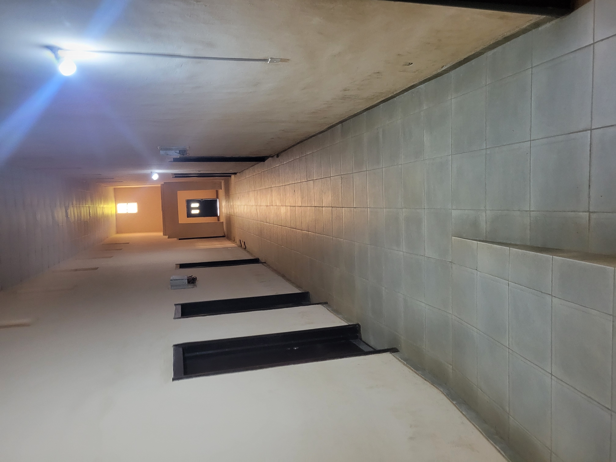 A long, narrow hallway with off-white walls and a tiled floor stretches into the distance. There are several dark-colored doors on the left side of the hallway, evenly spaced. On the right side, the wall appears to be textured and slightly discolored in places. A bright light fixture is visible on the right wall, casting a strong glare. The hallway leads to an area where a double door is visible, with a small, brightly lit window above it. The overall impression is of an interior corridor, possibly in a building with multiple rooms or apartments.