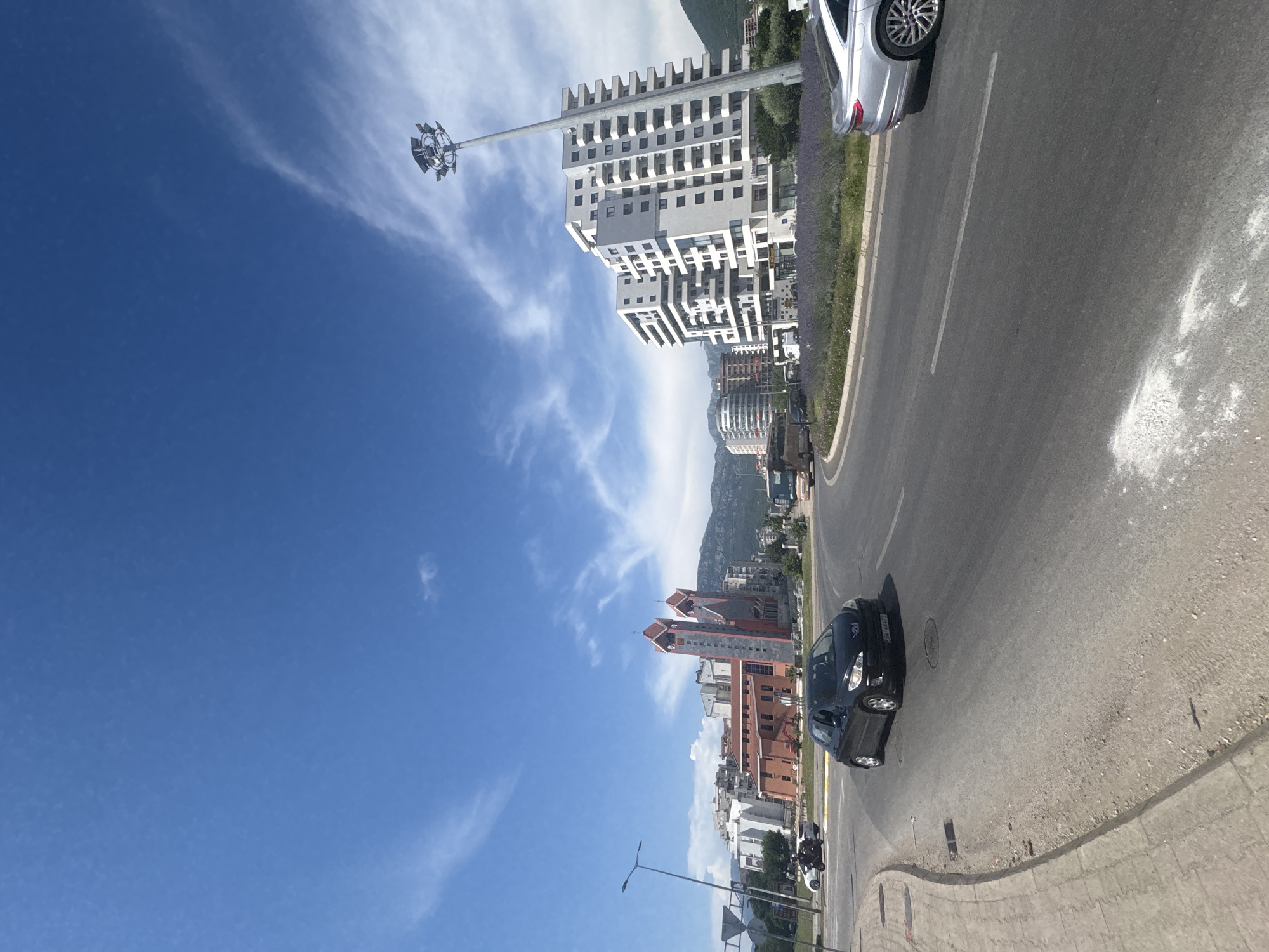 This is a street-level, wide-angle photograph of a modern city scene in Bar, Montenegro, on a clear, bright day with scattered white clouds in a blue sky. The perspective is from a road, likely a roundabout or a large intersection, looking towards a cluster of buildings.
In the foreground, a black Peugeot hatchback is driving on the asphalt road. To the right, a silver Audi sedan is also visible on the road. The road surface shows some wear and tear, with white markings and patches of lighter colored debris.
Mid-ground, to the left, a red brick building with two tall, distinct towers, possibly a church or a similar institutional building, stands prominently. Several other modern residential and commercial buildings, mostly white, are situated behind and around it. A street lamp with multiple fixtures is on the right side of the frame, near a tall, white, multi-story building with many balconies and square windows. A circular garden with purple flowers is located in front of this building.
In the background, more buildings and green, mountainous terrain can be seen, suggesting the city is nestled within a hilly or mountainous region. A bus is also visible in the distance, on the right side of the road. A white motorcycle with two riders is seen on the left side of the frame, near a traffic sign. The overall impression is one of urban development with a blend of older architectural styles (the red brick building) and modern construction.
