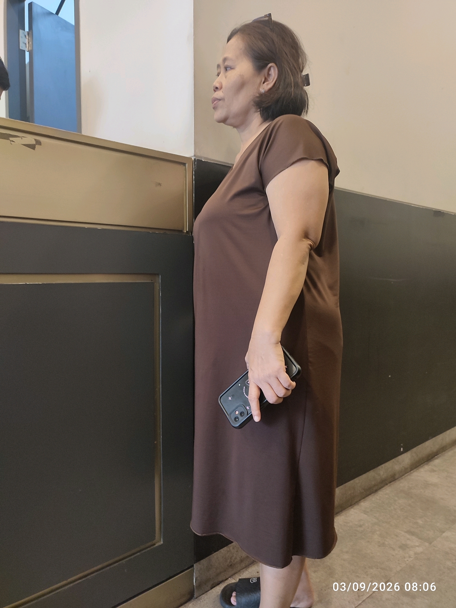A medium shot captures an East Asian woman in a brown dress standing in what appears to be an indoor public space, possibly a bank or government office, given the counter and muted color scheme. She is seen in profile, looking towards the left side of the frame. The woman has short, dark hair and is holding a dark-colored smartphone in her right hand, with her fingers around the device. Her left arm hangs loosely at her side. The background consists of a cream-colored wall and a dark gray panel with a golden trim running horizontally across the middle of the frame. The flooring is a light-colored tiled surface. A date and time stamp, "03/09/2026 08:06," is visible in the lower right corner of the image. The overall impression is that of a candid moment, possibly capturing the woman waiting or interacting at a service counter.