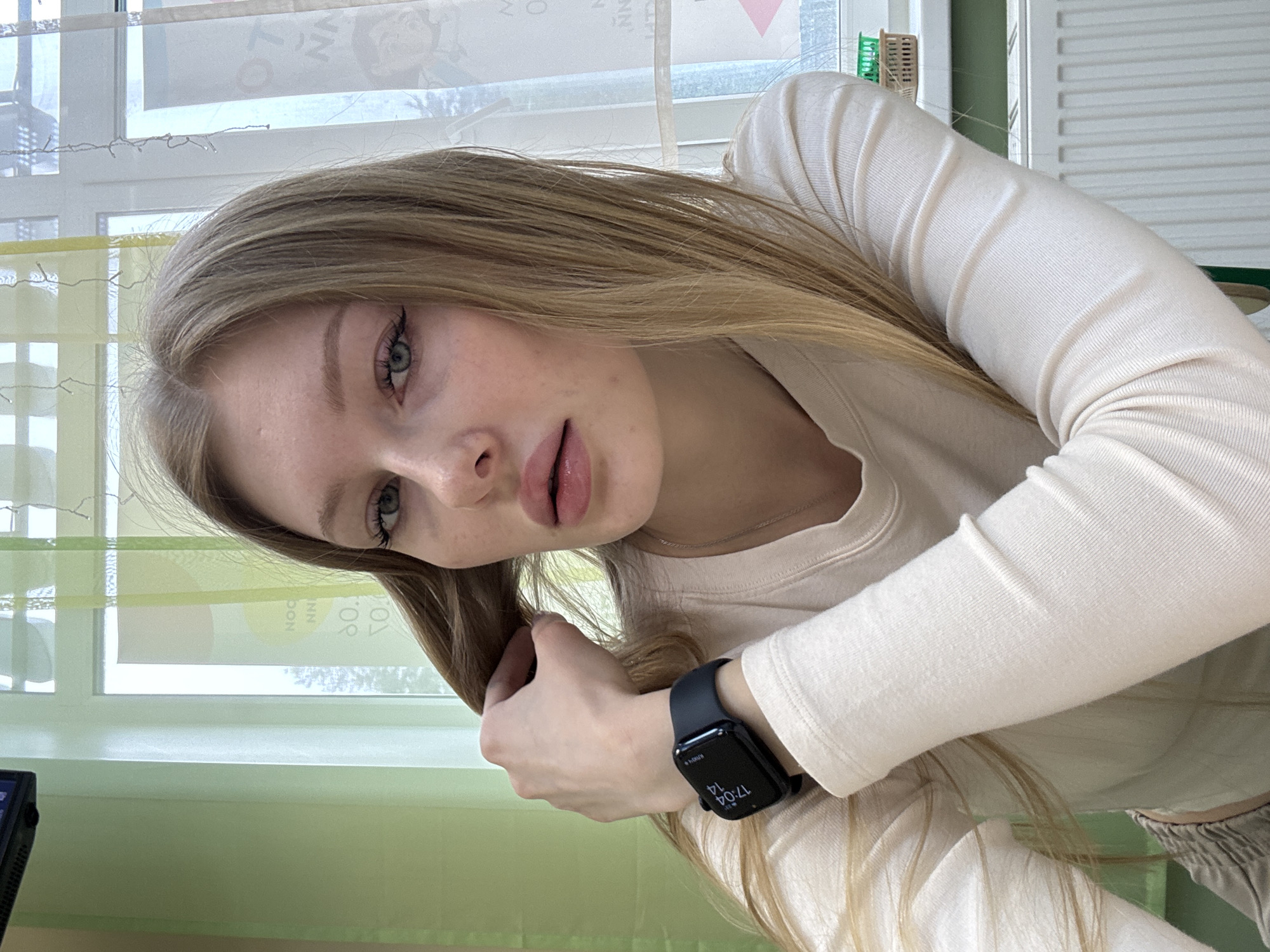 A young fair-skinned woman with long blonde hair is facing the camera indoors. She is wearing a long-sleeved light-colored shirt and has a black smartwatch on her left wrist. The smartwatch displays the time as 17:04. She is holding a strand of her hair. The background includes a window with blinds. Through the window, some elements indicate a possible location in Perm, Russia, but they are not definitively identifiable.