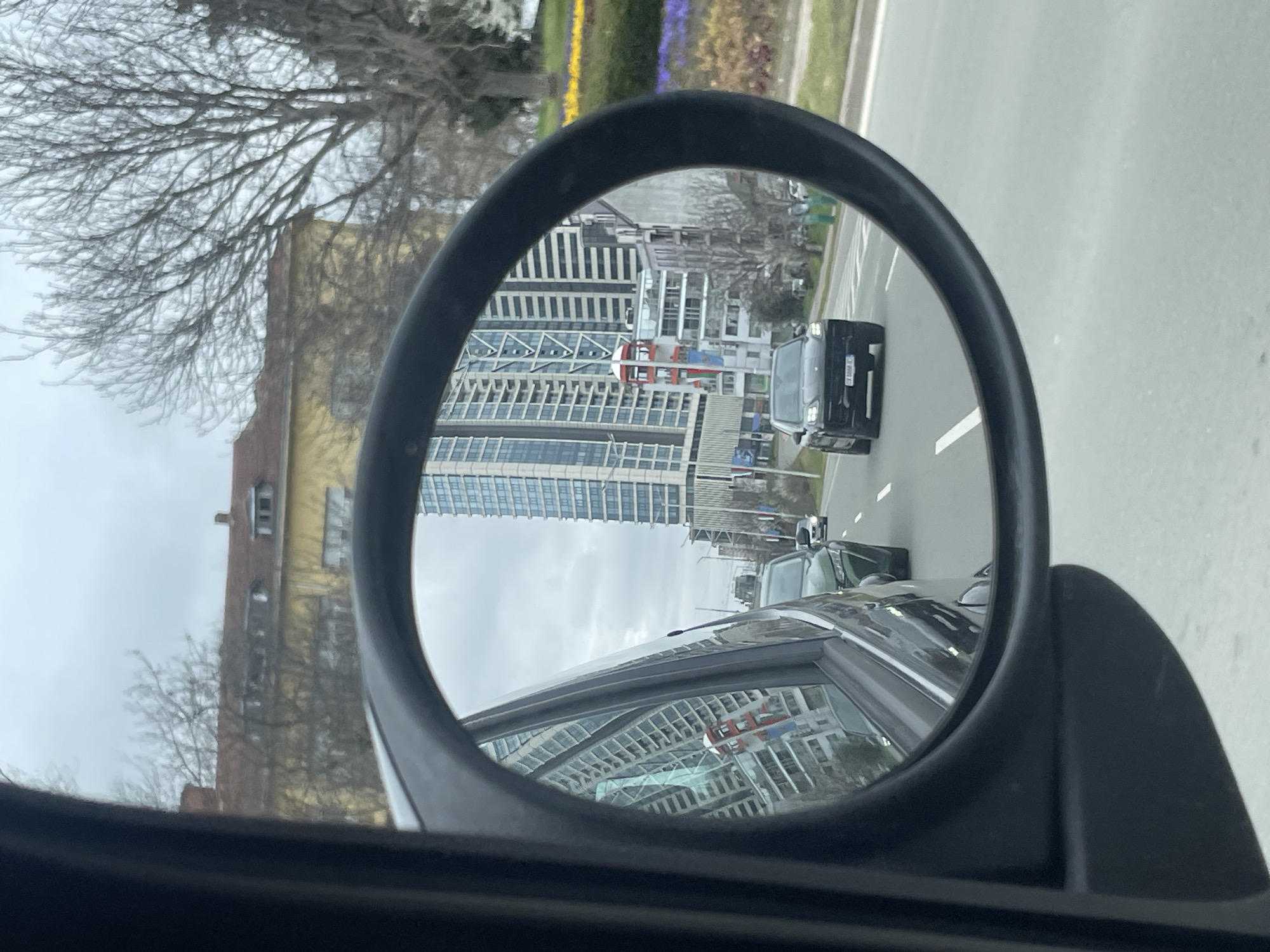 The image shows a car's side mirror reflecting a street scene in Sofia, Bulgaria. Visible in the mirror are several vehicles, including a black car directly behind the photographer's vehicle. High-rise buildings with distinctive architectural features are reflected, including a red and white structure. Outside the mirror's frame, to the left, is a building with yellow walls and bare trees in front of it. A street with a median with yellow and violet flowering plants can be seen on the right side. The sky is overcast.