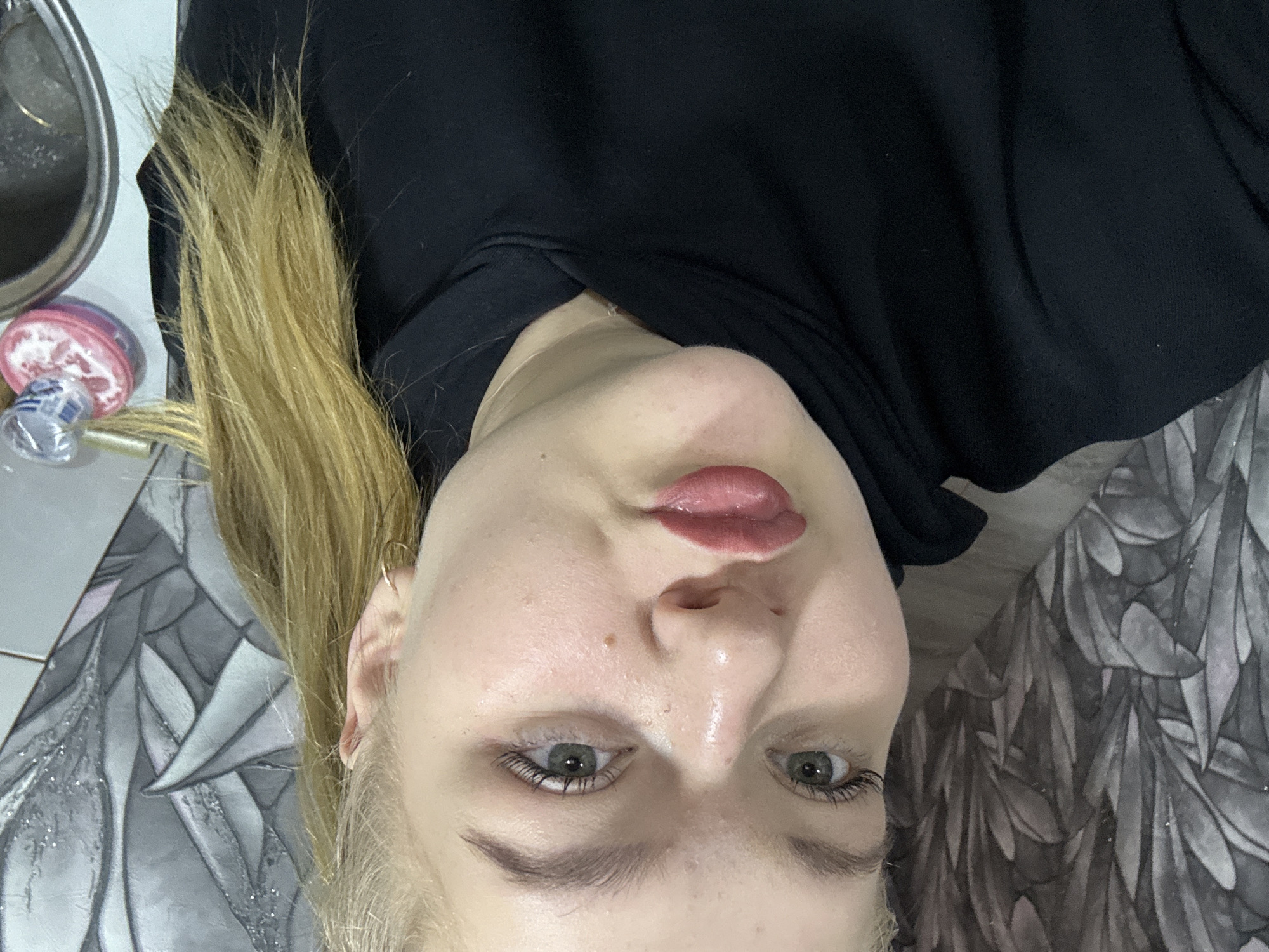 A close-up selfie portrait of a fair-skinned woman with blonde hair and green eyes. She is wearing a black hoodie. Her lips are pursed. The background includes a sink, toiletries, and wallpaper with a feather pattern. Location information is not discernible from the image.
