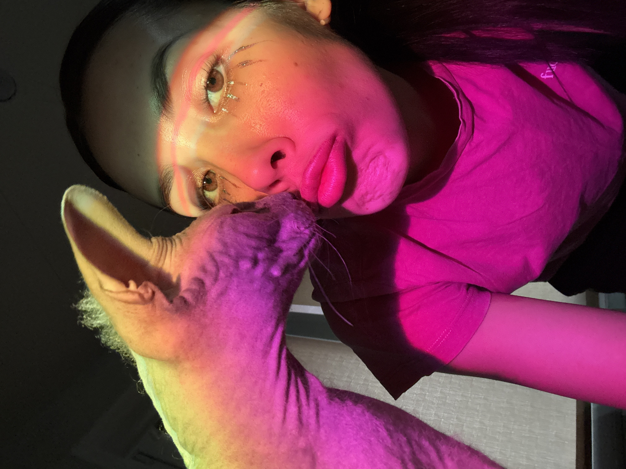 A woman with dark hair and light skin is looking at the camera. She is wearing a pink shirt and has small crystals under her eyes. A hairless cat is positioned near her face, possibly nuzzling her. The scene is illuminated by a pink and yellow light. The image does not include information that allows it to be located in Votkinsk, Russia.