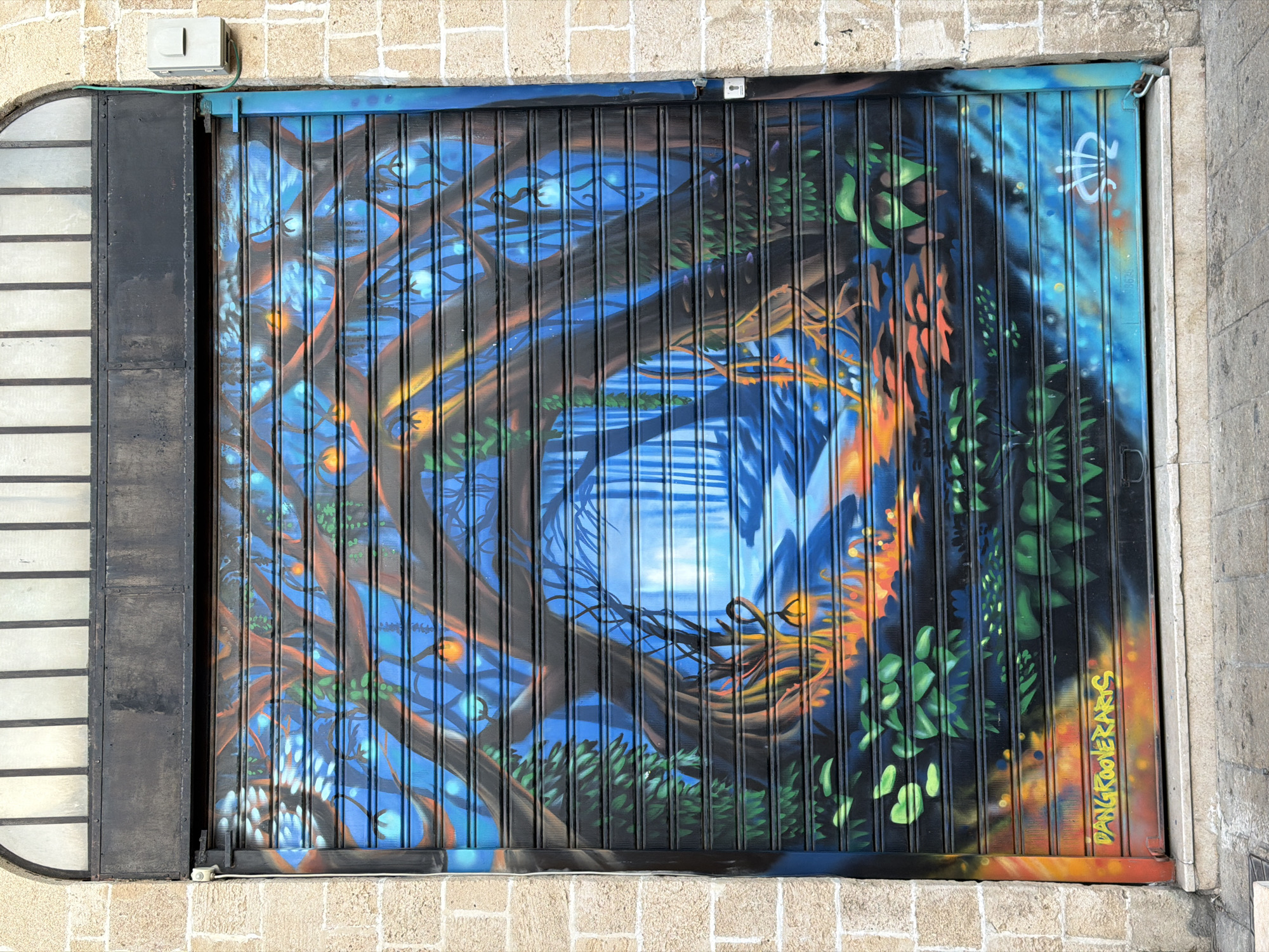The image shows a painted metal roll-up door in Tel-Aviv, Israel. The door features a surrealistic nature scene dominated by blues and greens, depicting stylized trees, foliage, and a body of water. The painting incorporates oranges and yellows, suggesting light sources within the scene. The lower right corner of the door has an artist's signature, "DANGKOOLERANKS." To the left of the painted door is a black metal frame and opaque glass or plastic pane. The door is set into a beige brick building. At the top left of the door is a white electrical box.