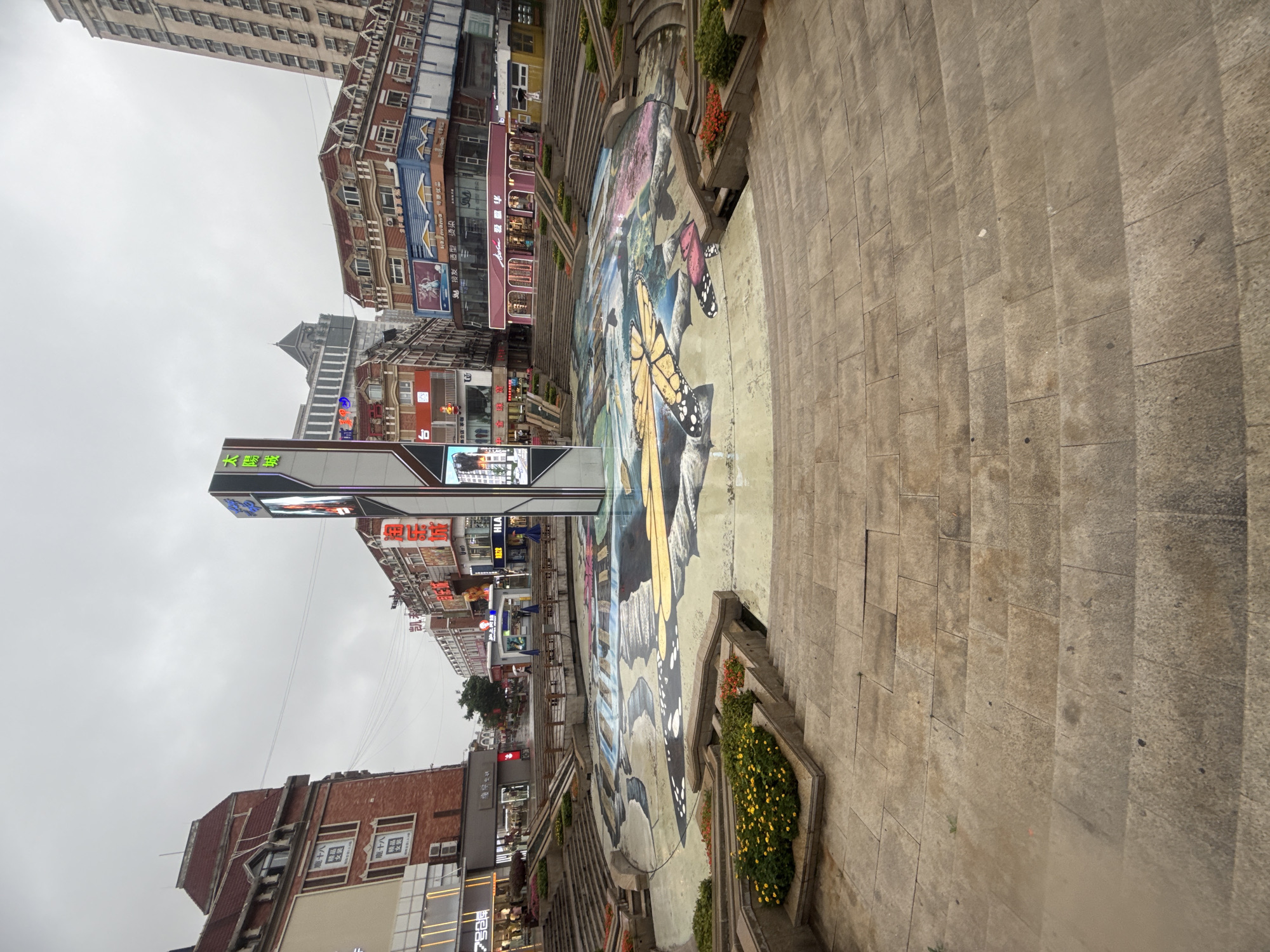 The image depicts a wide, multi-tiered plaza with a central, artistic fountain area. The plaza is paved with large, flat stone tiles, and tiered seating surrounds the fountain. The fountain itself is a large, shallow basin adorned with a colorful mural featuring oversized butterflies and abstract patterns. A tall, modern, multi-sided digital display stands at the center of the fountain, showing advertisements and graphics.
Surrounding the plaza are several multi-story buildings with diverse architectural styles. Some buildings exhibit traditional European-inspired designs with red tile roofs and ornate facades, while others are more modern, featuring large windows and prominent signage. The buildings house various businesses, indicated by illuminated signs in Chinese characters, some of which are neon. Examples of businesses visible include clothing stores and what appears to be a shopping mall.
The sky is overcast, suggesting a cloudy or possibly rainy day, which may have contributed to the wet appearance of the stone paving. A few people are visible in the distance, walking through the plaza and near the buildings, some holding umbrellas. The overall scene suggests a public urban space, likely a commercial or entertainment district, located in Qinhuangdao, China. The specific location is identified as the intersection of Wenhua Road and Dalian Road in Haigang District, Qinhuangdao.