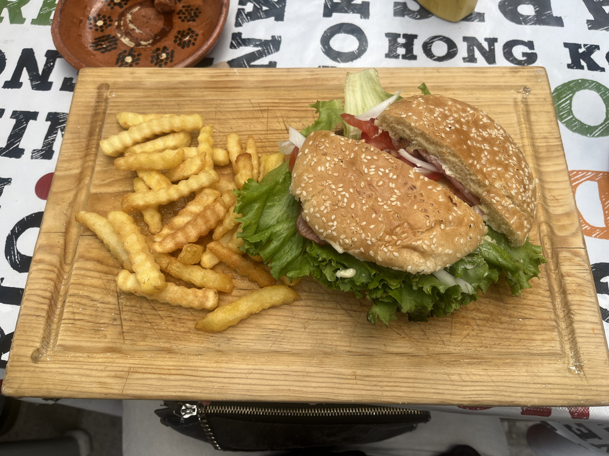 On a light wooden cutting board sits a hamburger cut in half and a pile of french fries. The burger is on the right side of the board, and the fries are on the left. The burger has sesame seeds on the bun and is filled with lettuce, tomato, onion, and an indiscernible sauce. The fries are a medium yellow color.
The background is a patterned tablecloth. Above the cutting board is a brown bowl with black dots arranged in a floral pattern.
The cutting board and food are situated in San Francisco Totimehuacán, Mexico.