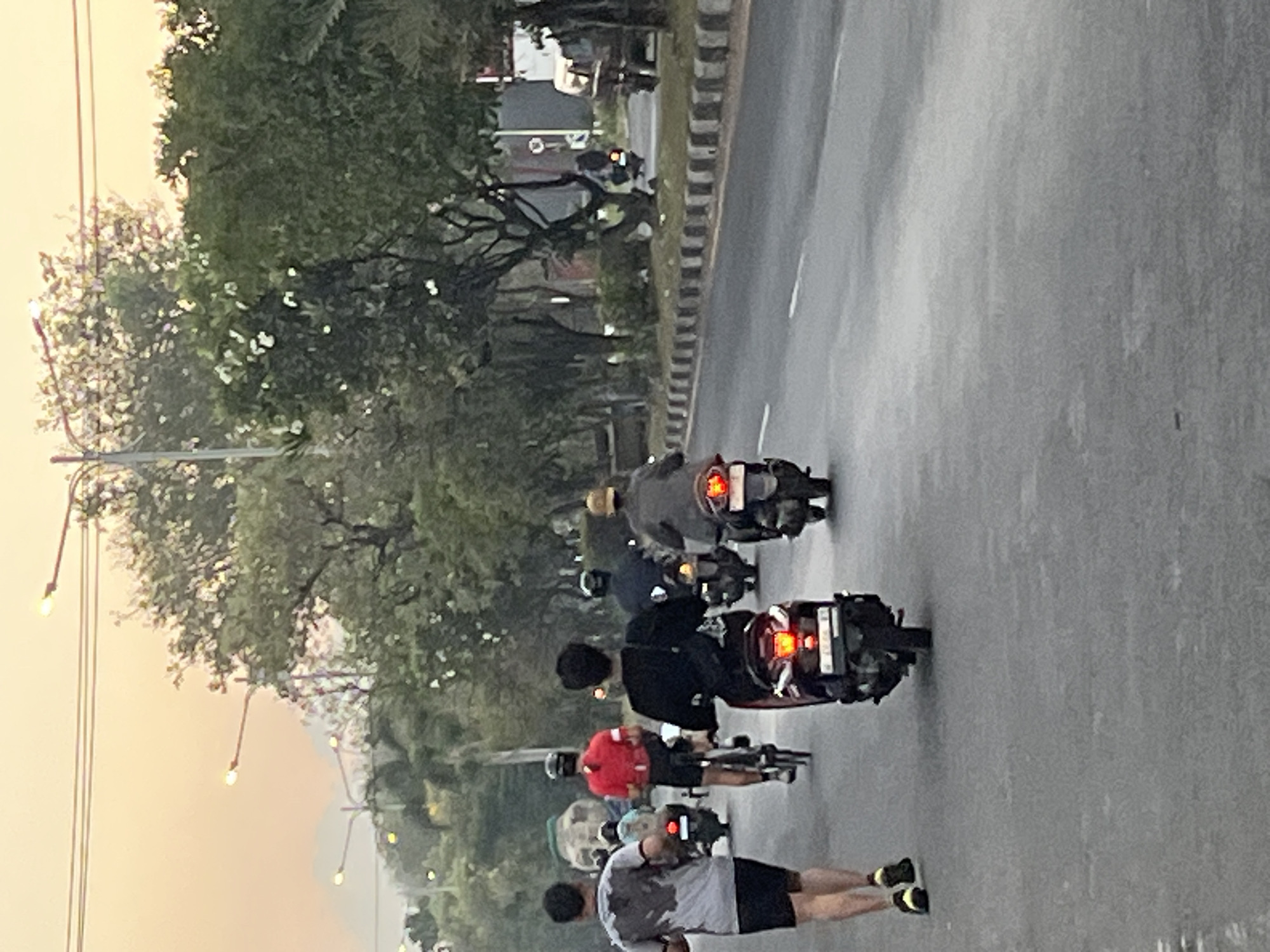 A group of people are on a street in Boro, Indonesia, during what appears to be dawn or dusk. The sky has a warm, orange hue. Several individuals are engaged in activities: a person in a grey shirt and black shorts is running on the left side of the road. Next to them, a person in a red shirt and black shorts is riding a bicycle. Further down the road, a person in a black shirt is riding a scooter, followed by another person in a dark outfit on a scooter, and a person in a grey jacket and tan hat also on a scooter. Trees line the right side of the road, with some streetlights visible in the background. The road has a curb with black and white markings. A few other vehicles, including a truck and a scooter, are visible in the distance on the right side of the road. The overall scene depicts a common street in an Indonesian city with various modes of transportation and pedestrian activity.