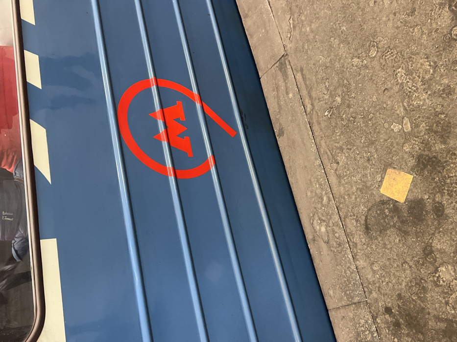 A blue subway train car with a red metro logo is seen next to a concrete platform. The logo features a stylized 'M' inside a circular design with an extended tail. Ribbed panels run vertically along the train car's surface. A small, yellow, square object is resting on the grey concrete platform. The lighting is bright, casting subtle shadows on the ground. The view is an angled, close-up perspective.